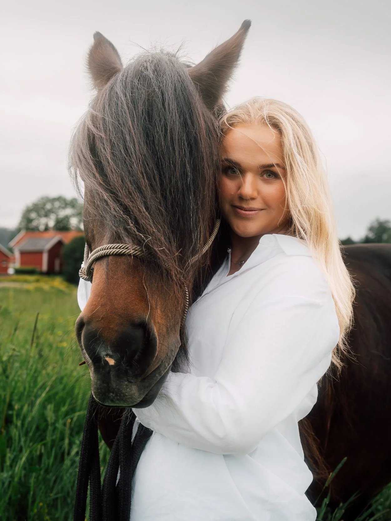 En kvinne med blond hår holder rundt en stor brunhvit hest i en grønn åker med hus i bakgrunnen.