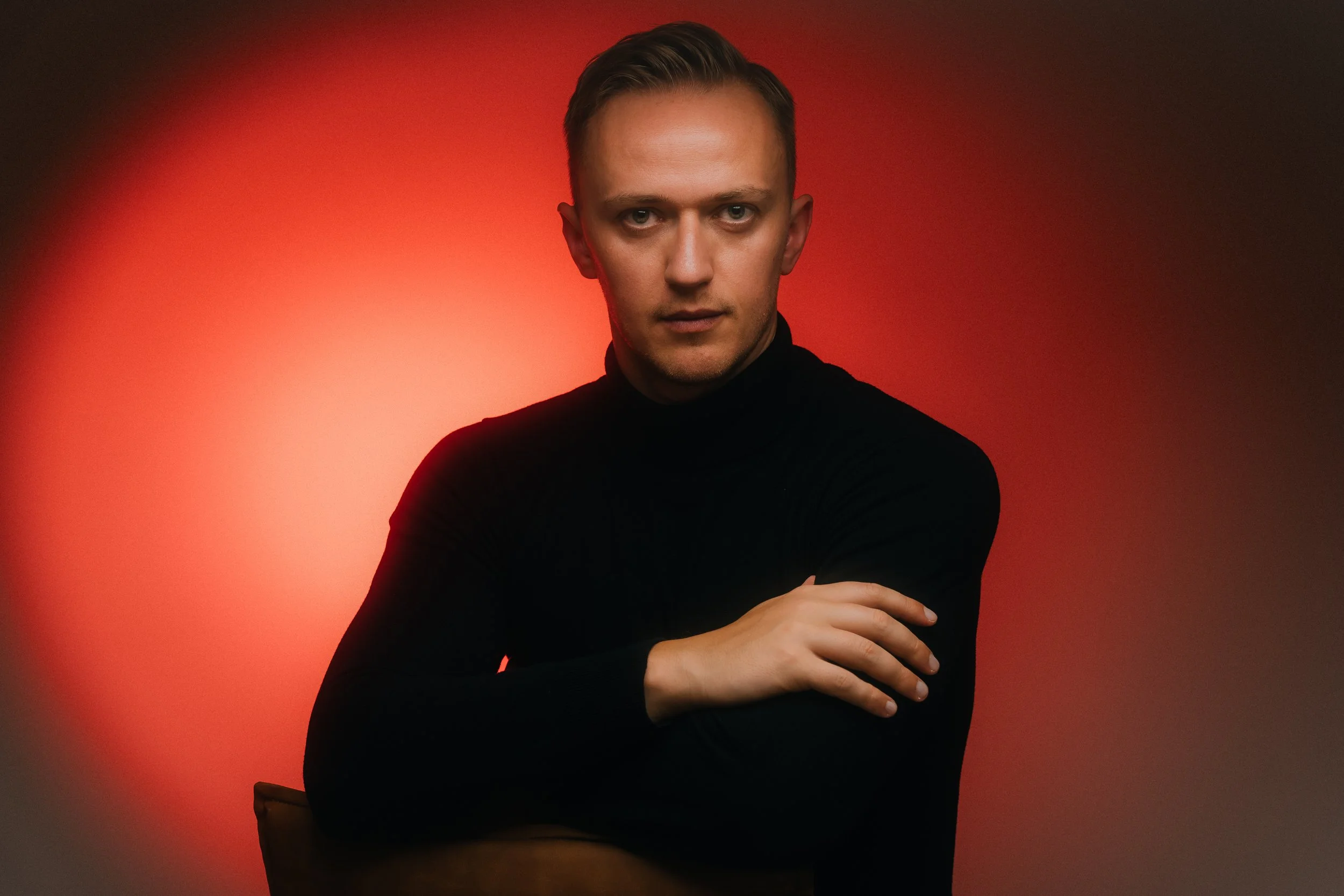 Young man with short, neatly styled hair posing with arms crossed in front of a warm red gradient background.