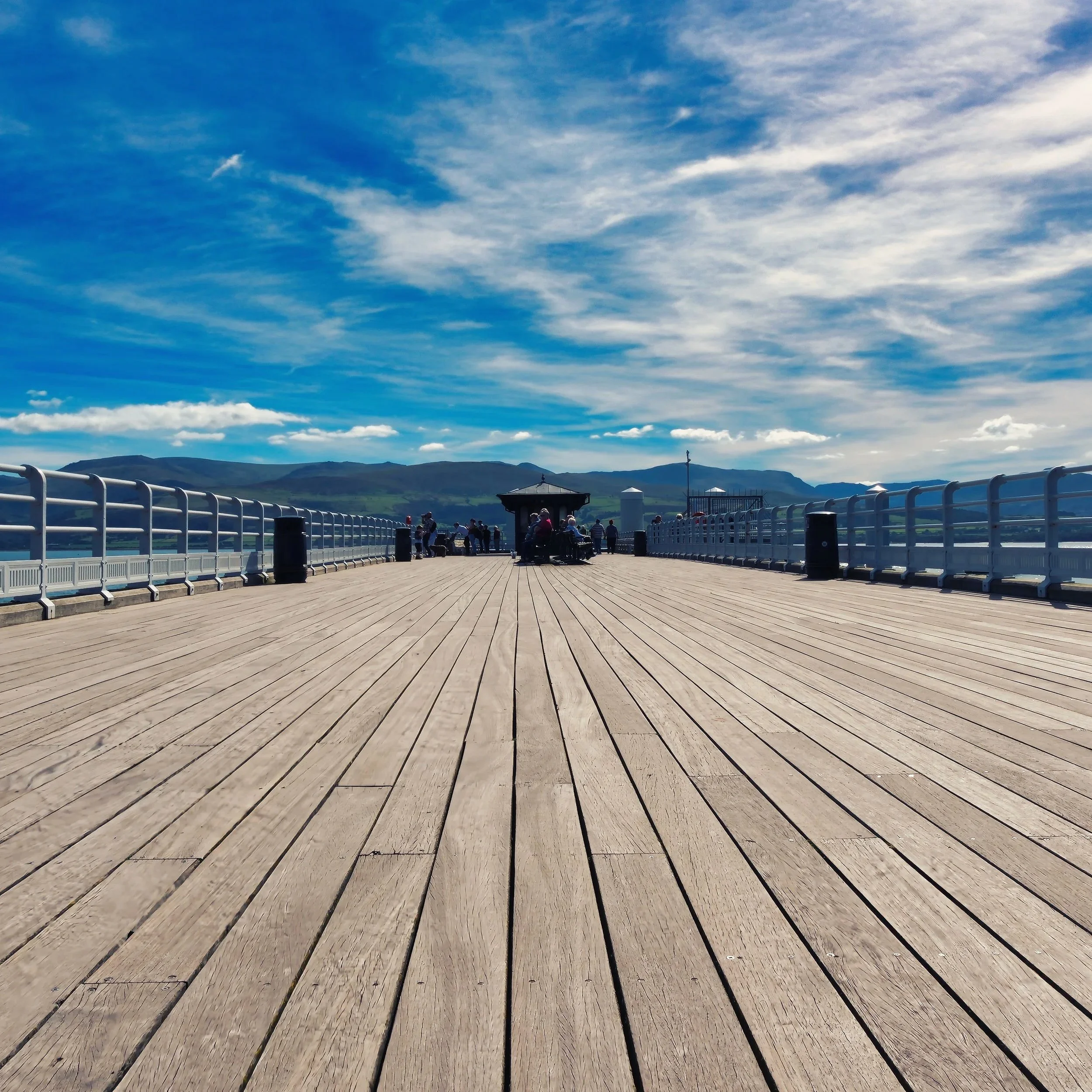 "Beaumaris Pier" Anglesey. A perspective piece, detailing the long wooden boards of Beamuris Pier, directing your attention to the  distant mountain range of Snowdonia. The mottled blue sky and scattered clouds frame the story