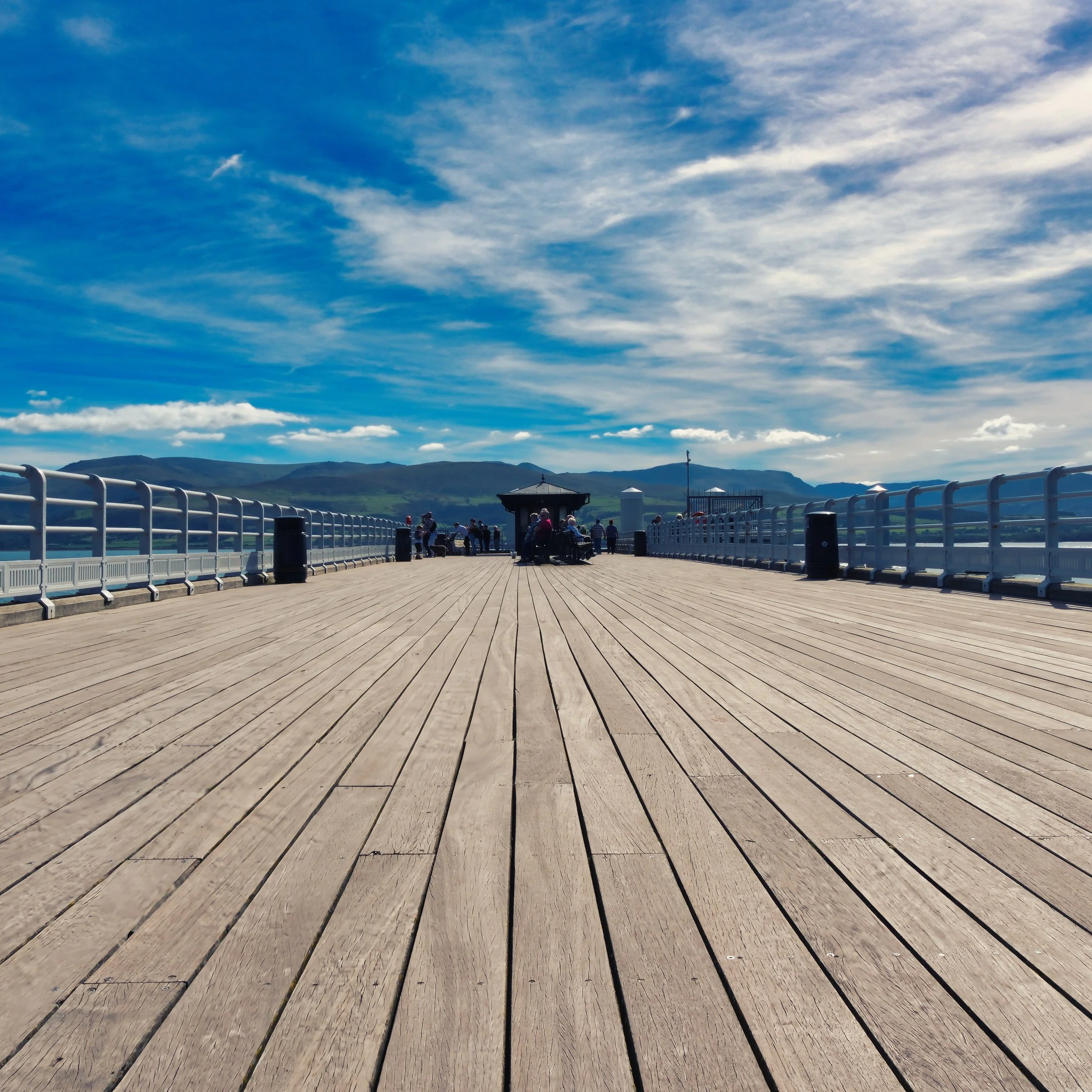 "Beaumaris Pier" Anglesea. A perspective piece, detailing the long wooden boards of Beamuris Pier, directing your attention to the  distant mountain range of Snowdonia. The mottled blue sky and scattered clouds frame the story 