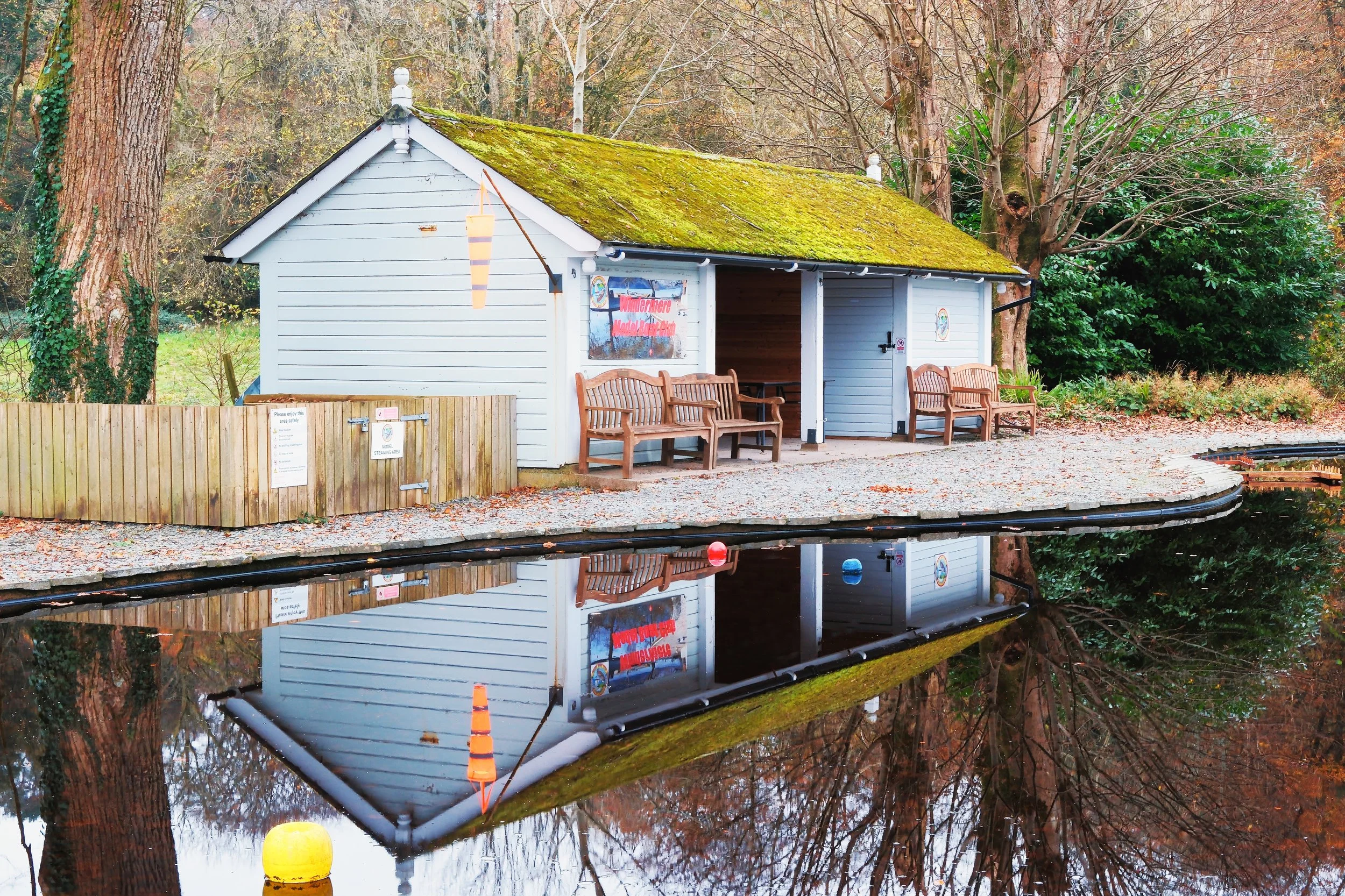 A small white wooden building with moss-covered roof and wooden benches outside, reflected in a calm body of water, surrounded by trees and autumn foliage.