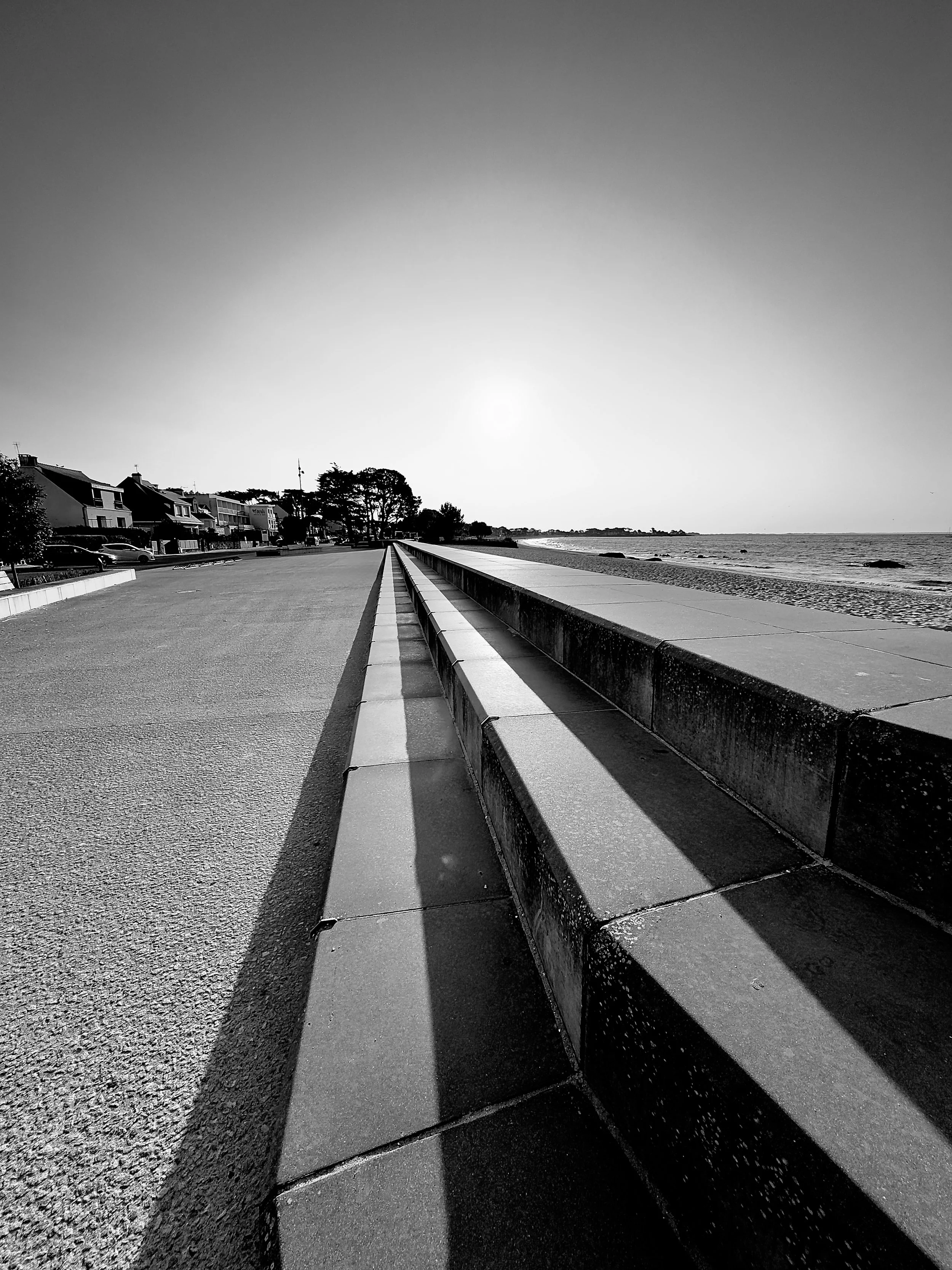 A black and white photo of a seaside walkway with houses on the left and the ocean on the right, featuring a long shadow cast across the pavement.