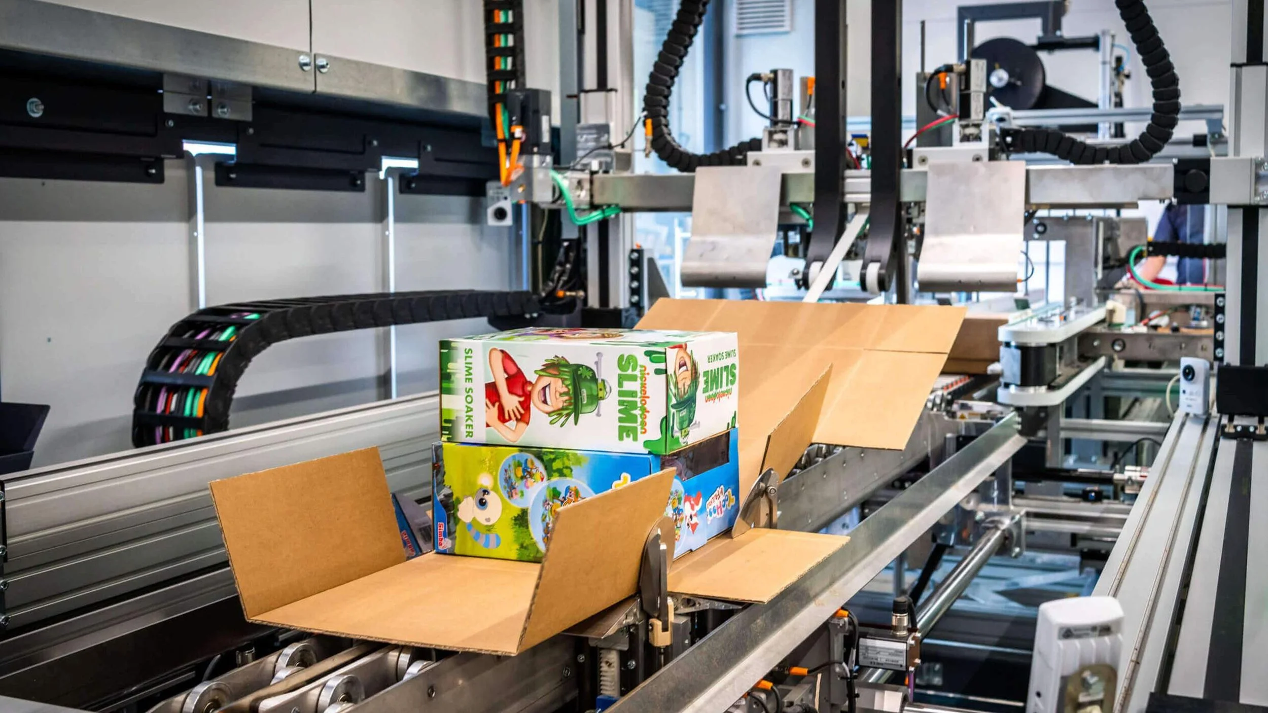 Conveyor belt with colorful toy boxes and an open cardboard box in a manufacturing or packaging facility.