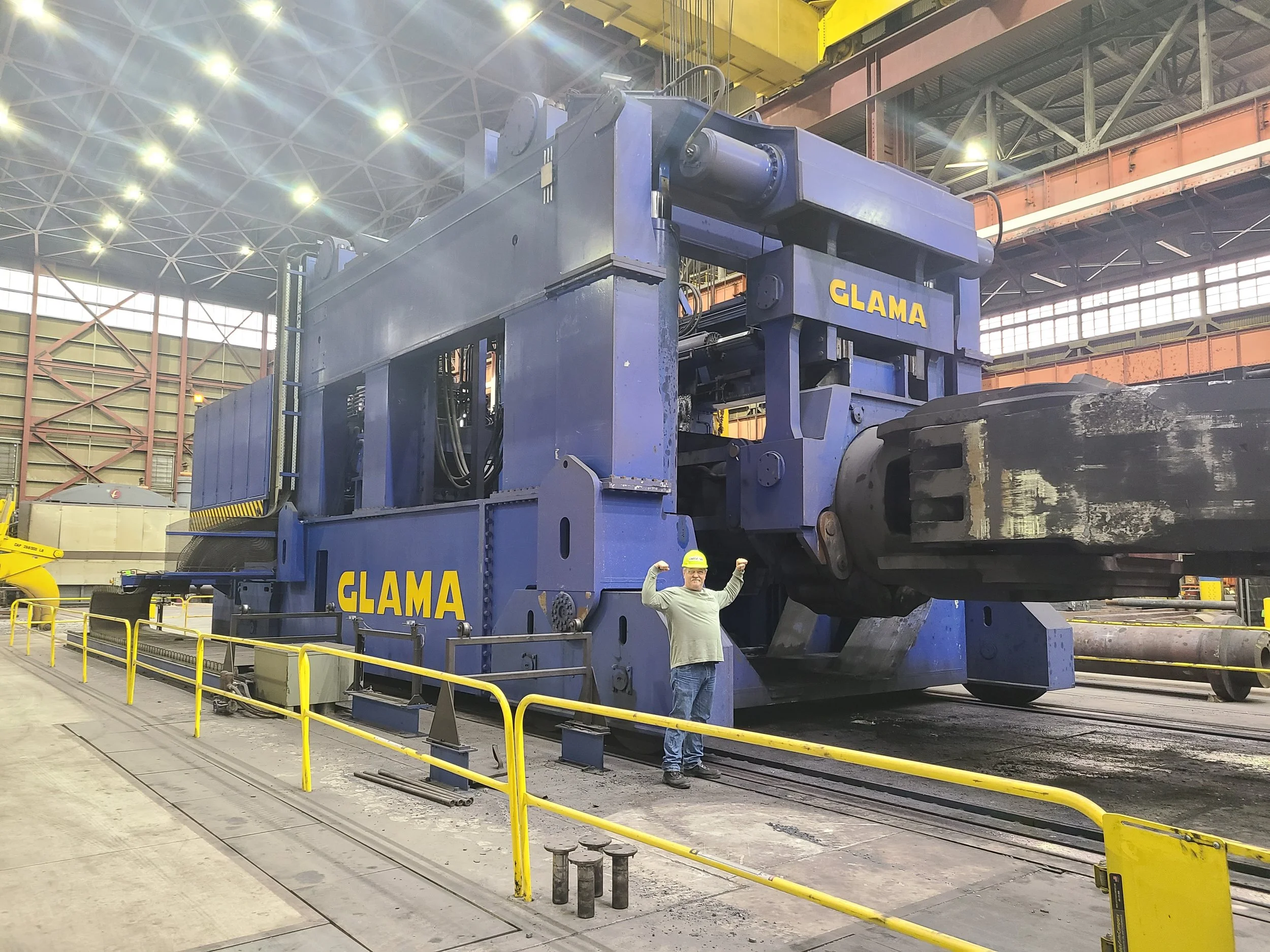 A large, blue industrial machine inside a factory or warehouse with yellow safety barriers, a worker in a white shirt and yellow helmet in front of it, and a man in a white shirt flexing his arms.