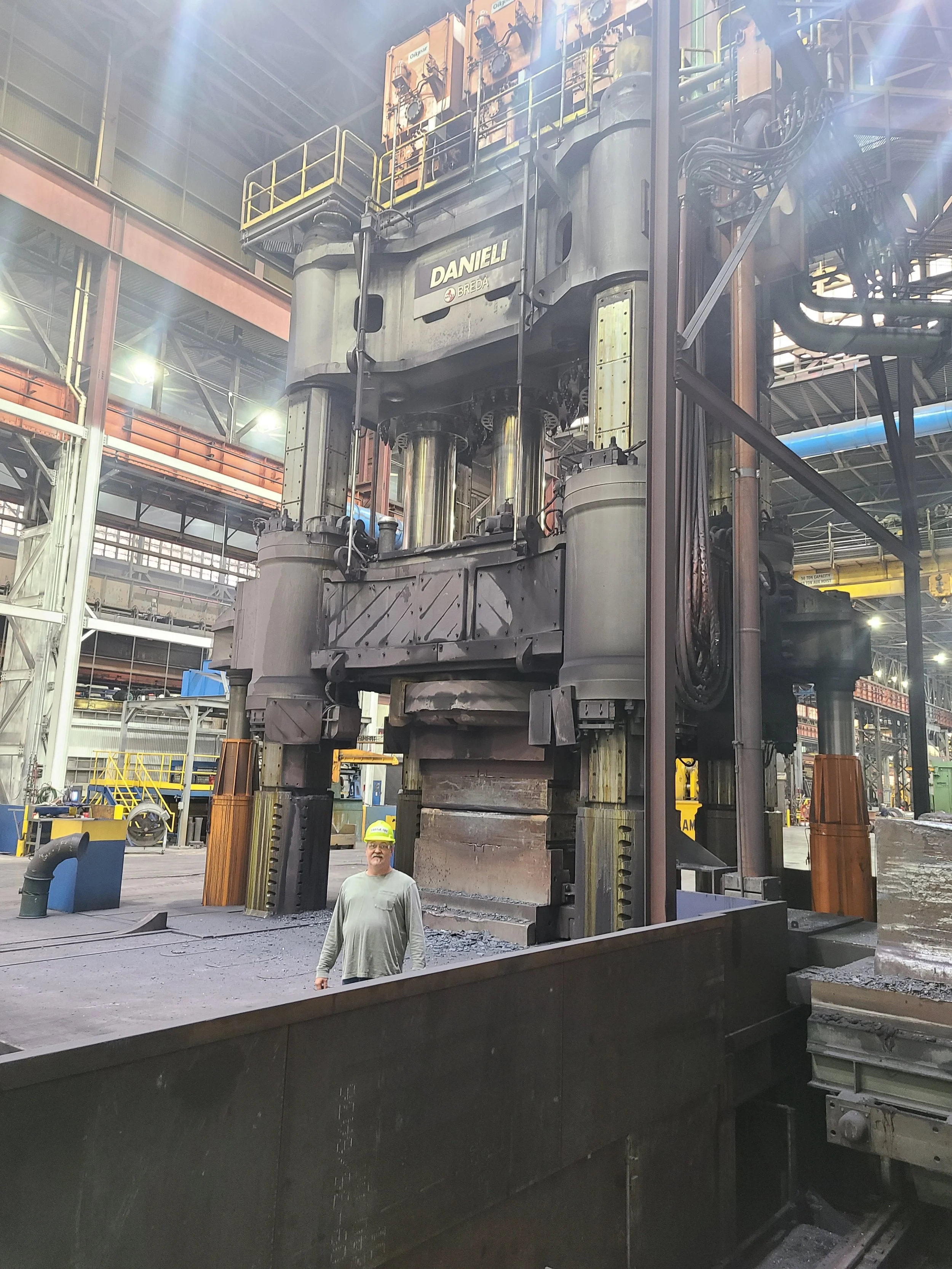 Large industrial machine in a factory setting with a person standing in front of it. The machine is gray with yellow safety rails on top and has multiple large metallic cylinders. Steel beams and various industrial equipment surround the scene.