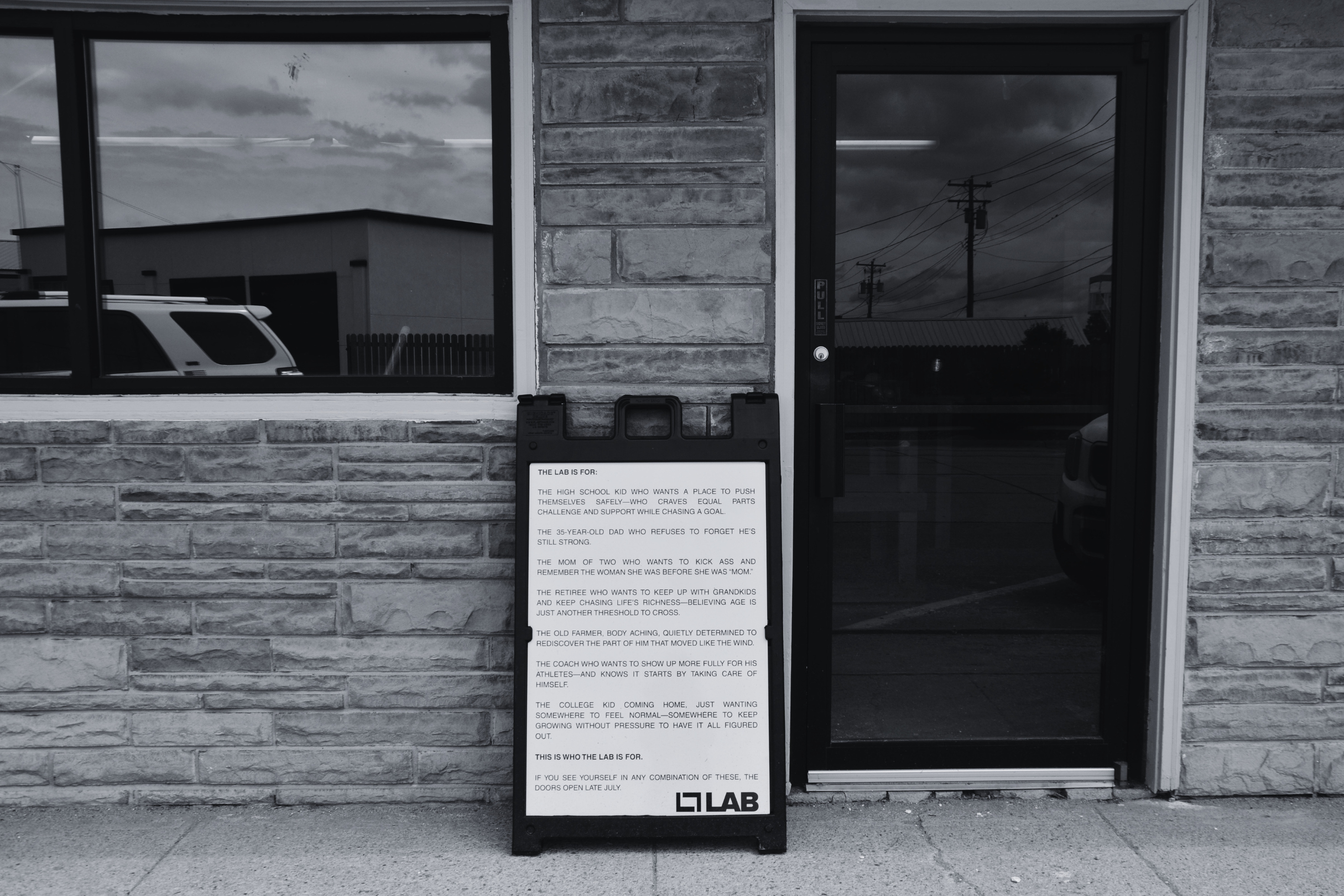 A black-and-white photo of a building exterior with two large windows and a door. There is a signboard in front with text explaining the purpose of a lab, and a logo at the bottom that reads 'L7 LAB'.