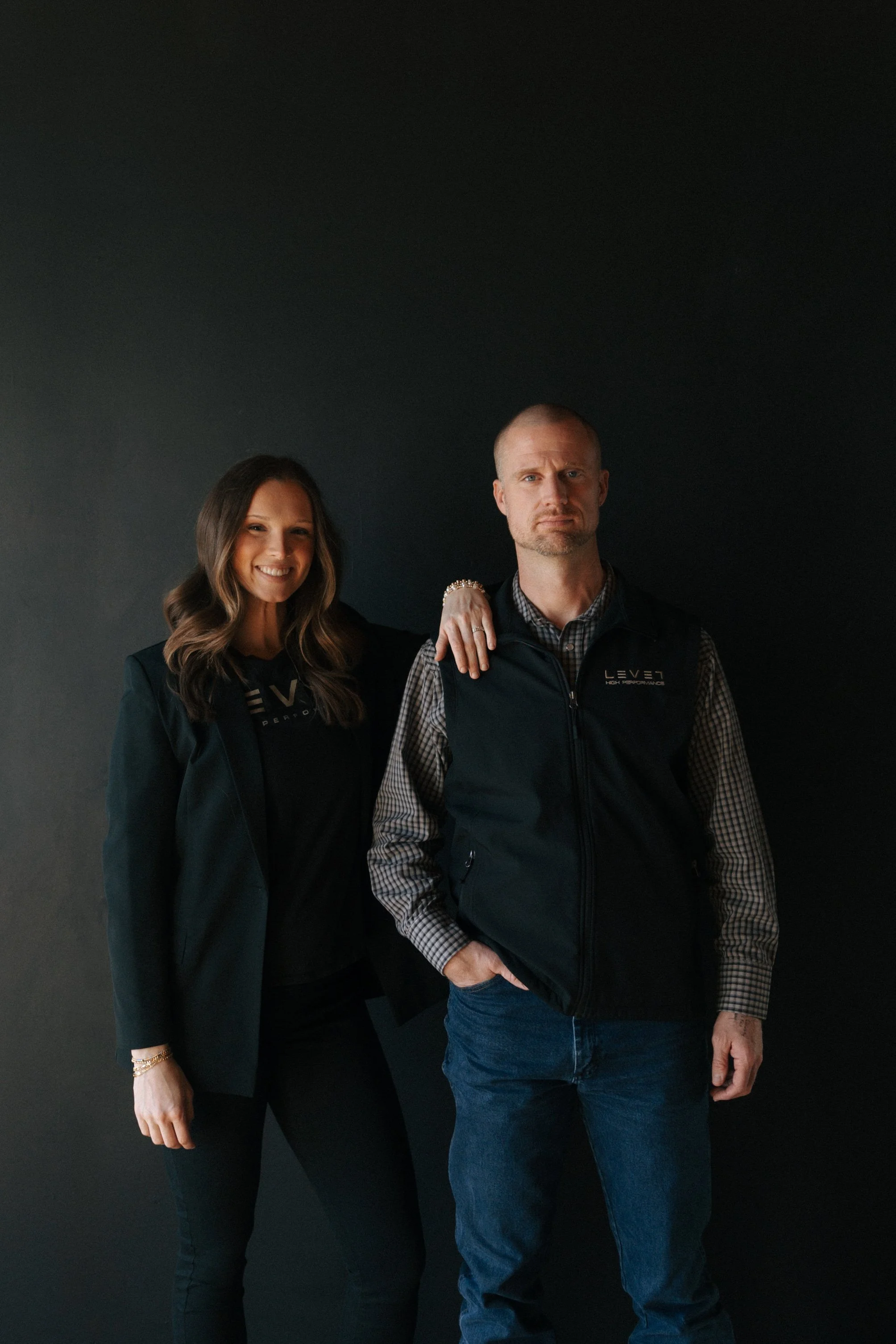 A woman and a man standing against a dark background. The woman has long brown hair, is smiling, and wears a black shirt with a green jacket. The man has a short beard, is serious, has his arm around the woman's shoulder, and wears a checkered shirt with a black vest that has a logo on it.