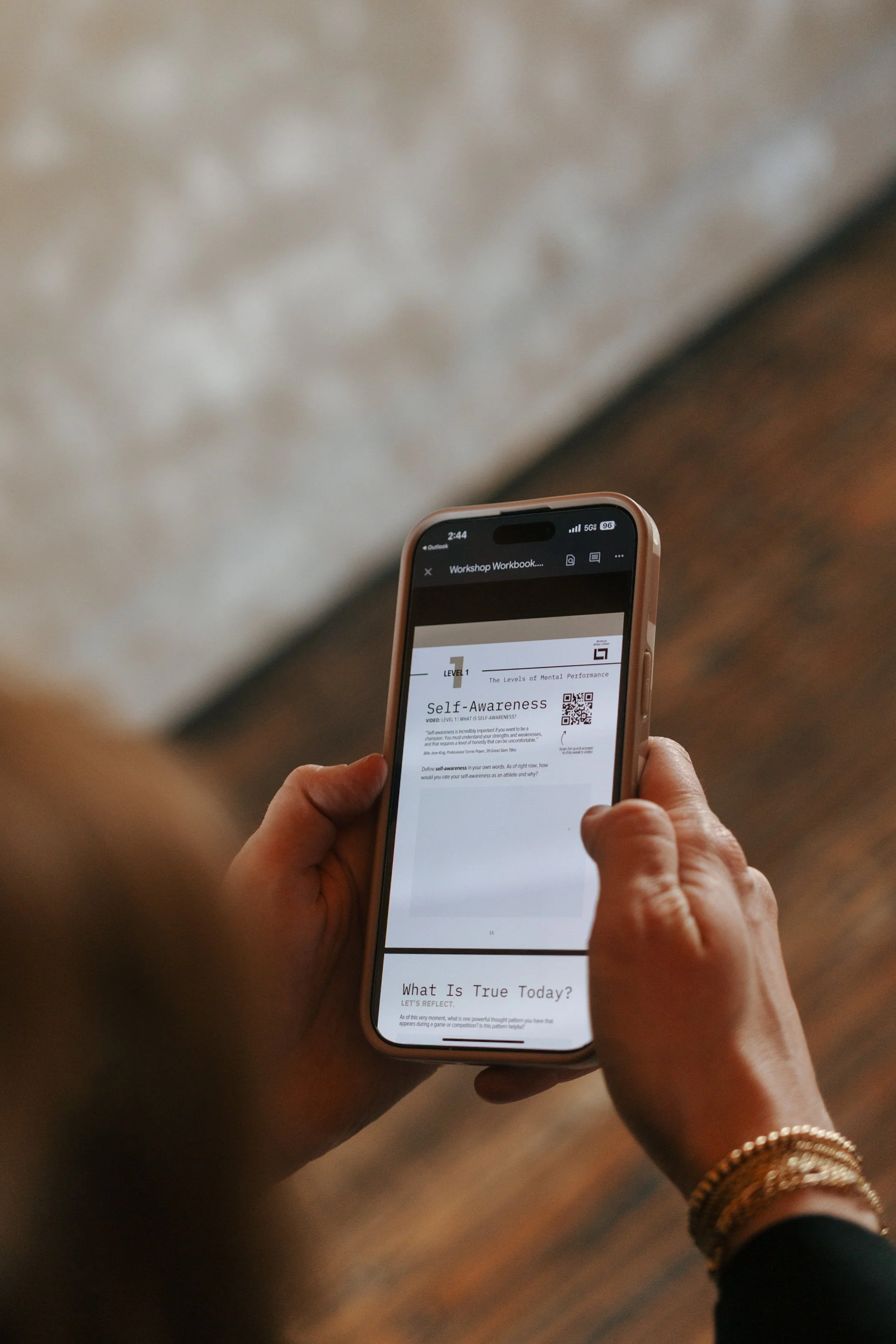 Person holding a smartphone displaying a workbook page titled 'Self-Awareness' with the subtitle 'What Is True Today?'. The person is wearing jewelry, including a gold bracelet, and is standing on a wooden floor.