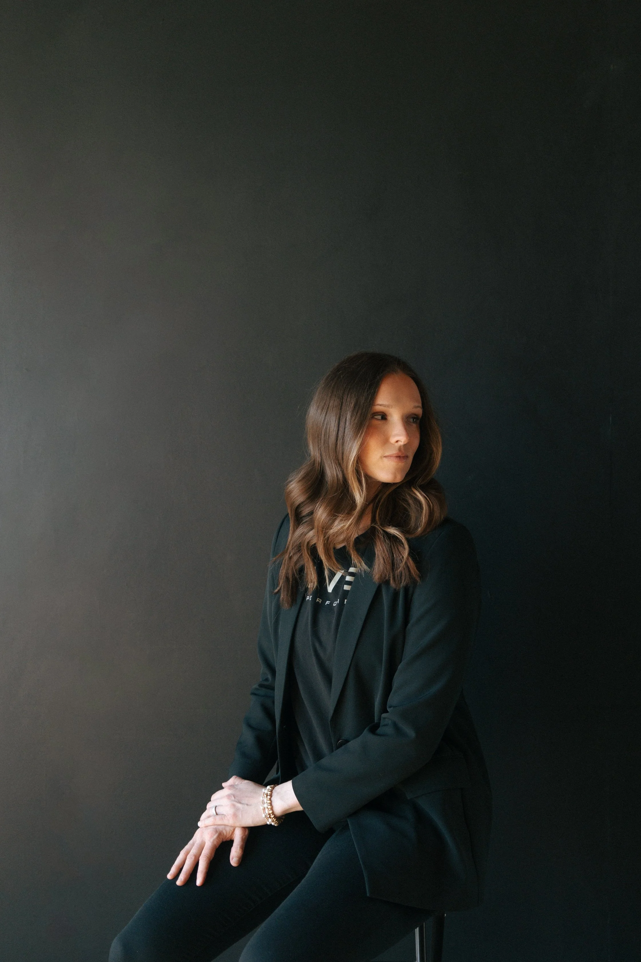 A woman with long wavy brown hair, wearing a black blazer and black pants, sitting against a dark gray background. She has jewelry on her wrist and fingers, and her head is turned slightly to the side with a neutral expression.