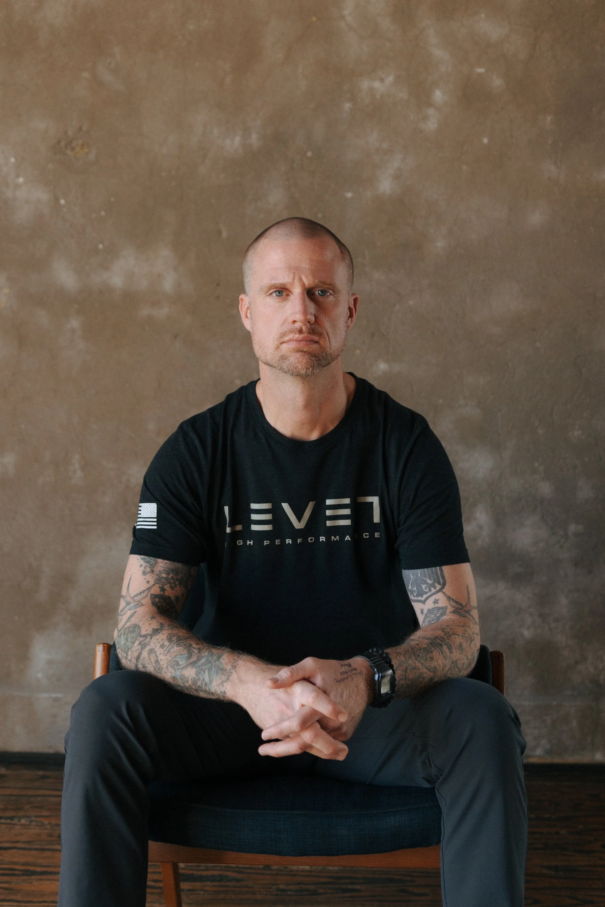 A man with tattoos on his arms, wearing a black t-shirt with the word 'LEVEL' on it, sitting on a chair in front of a textured beige wall with an intense expression.