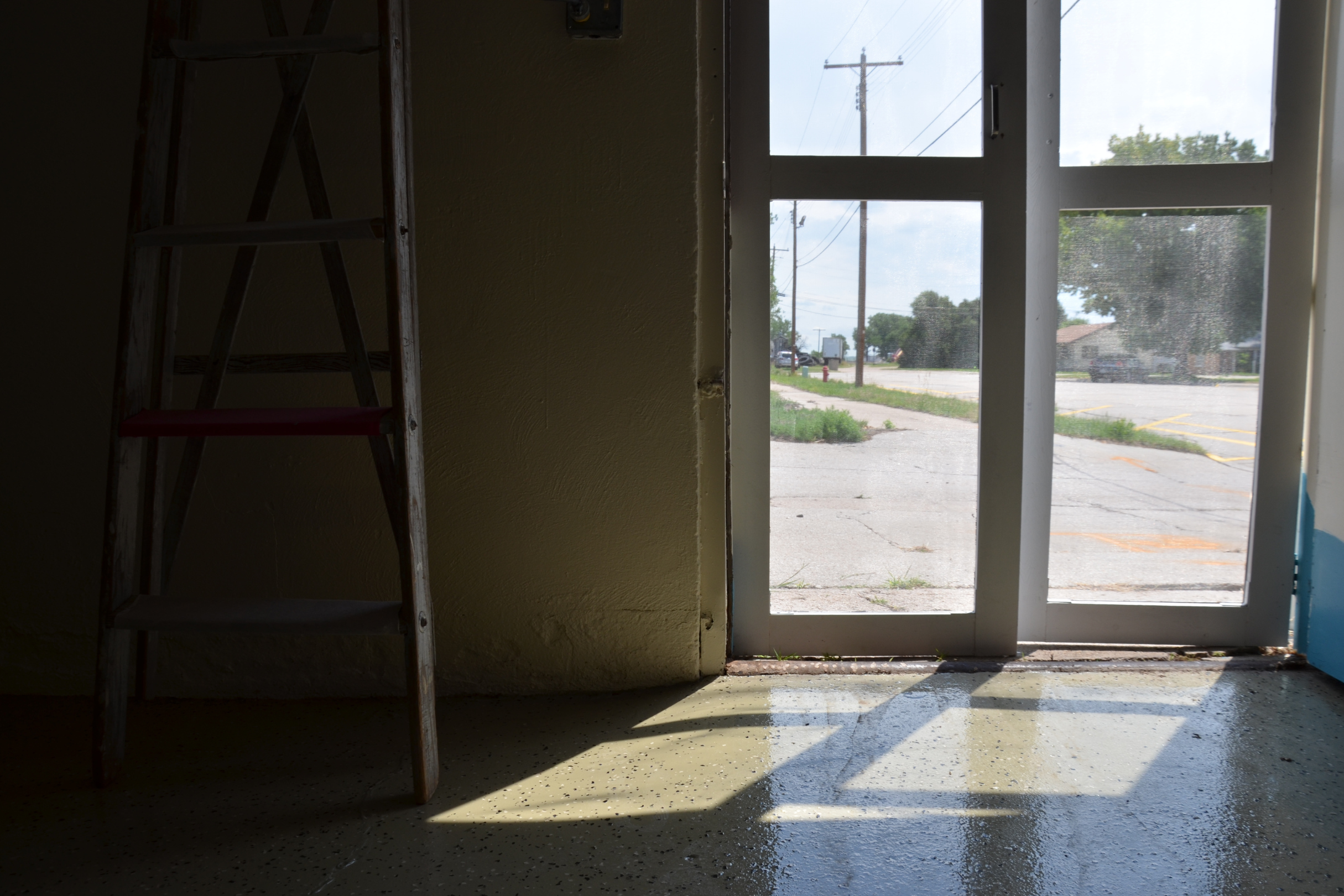 Inside view of a building looking out through a glass door, with sunlight casting a shadow on the floor; outside, a parking lot with some trees, utility poles, and a few buildings under a cloudy sky.