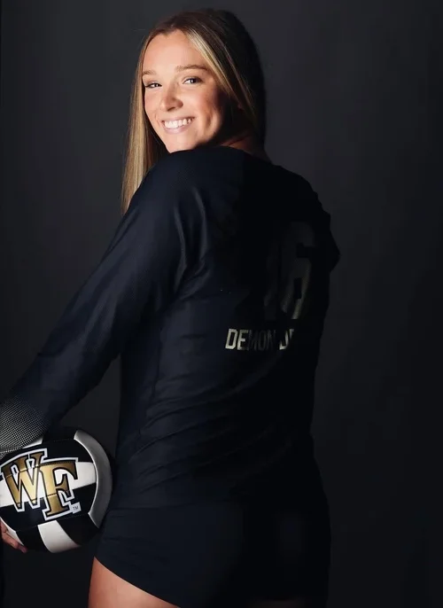 A young woman with long hair smiling and wearing a black athletic uniform holding a football with a black and gold design on it.