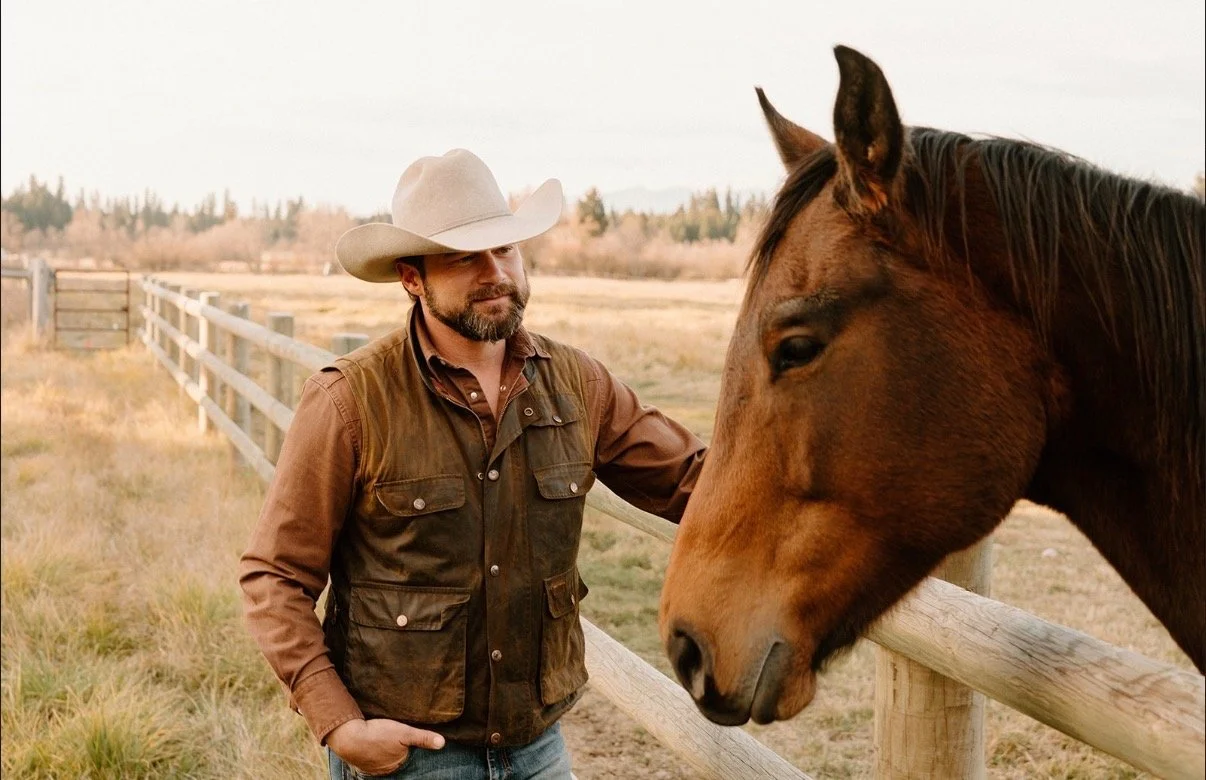 A man wearing a cowboy hat and brown vest petting a brown horse in a field with a wooden fence, trees, and mountains in the background.