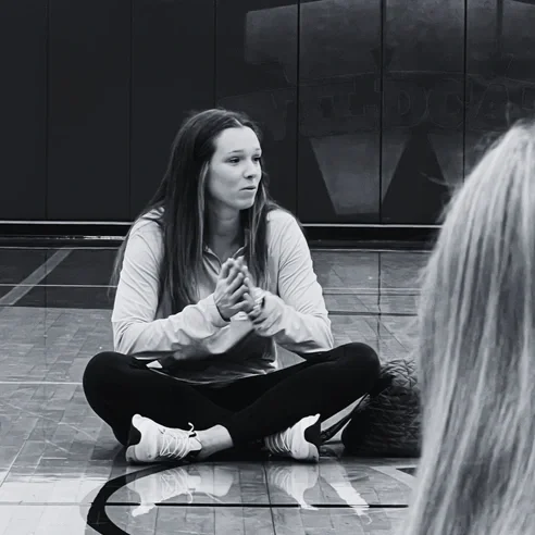 A young woman sitting cross-legged on a gym floor, clasping her hands in front of her, with a serious expression, in a gymnasium with a dark wall in the background.