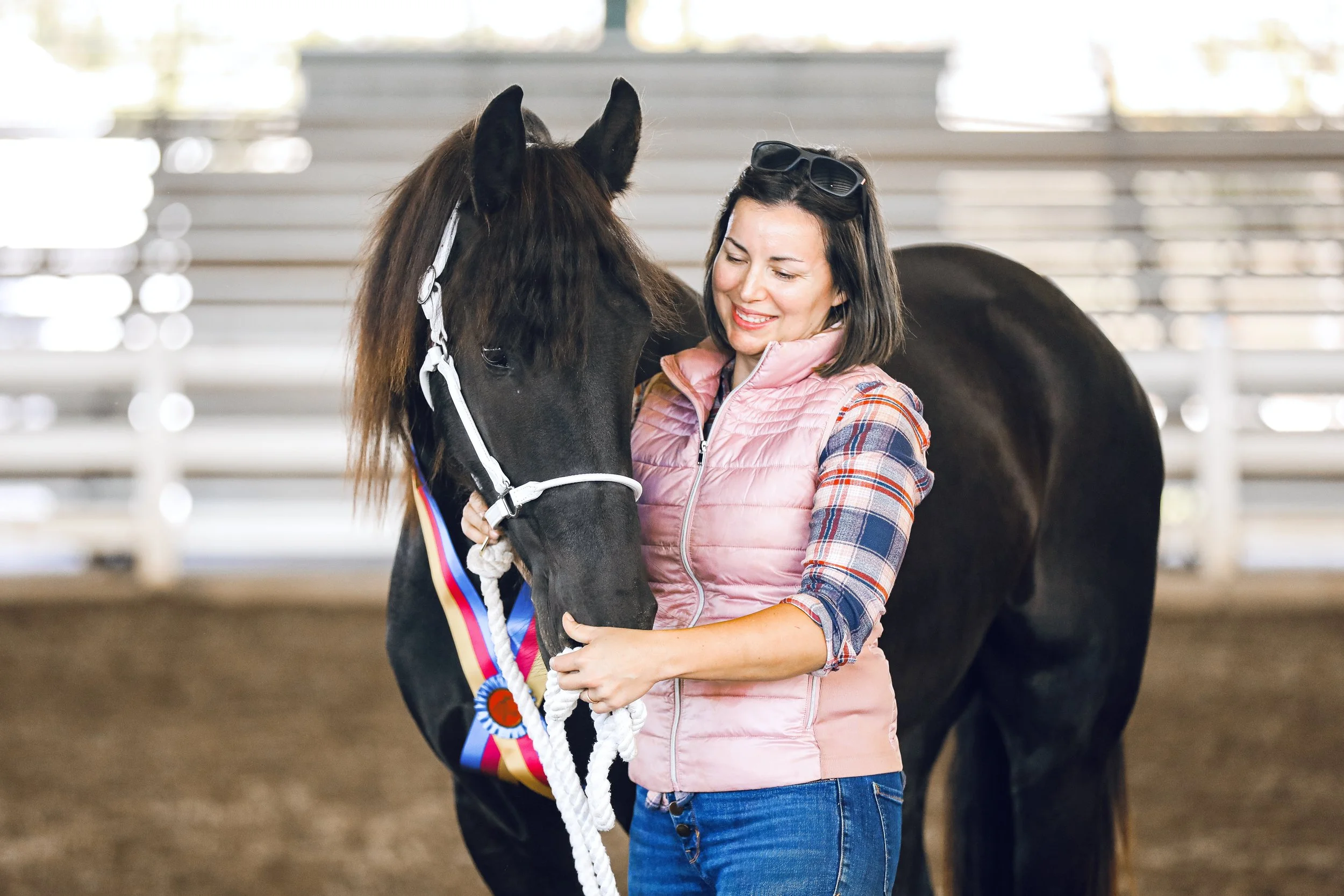 A woman smiling and hugging a black horse decorated with a ribbon and a rainbow-colored ribbon at a stable or barn.