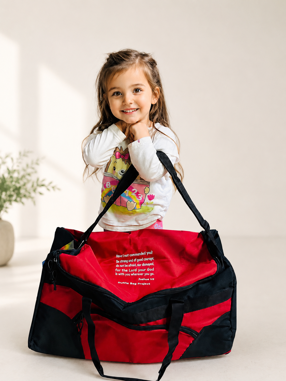Young girl with long brown hair and a big smile, standing behind a large red and black duffle bag, in a bright room with a light-colored background.