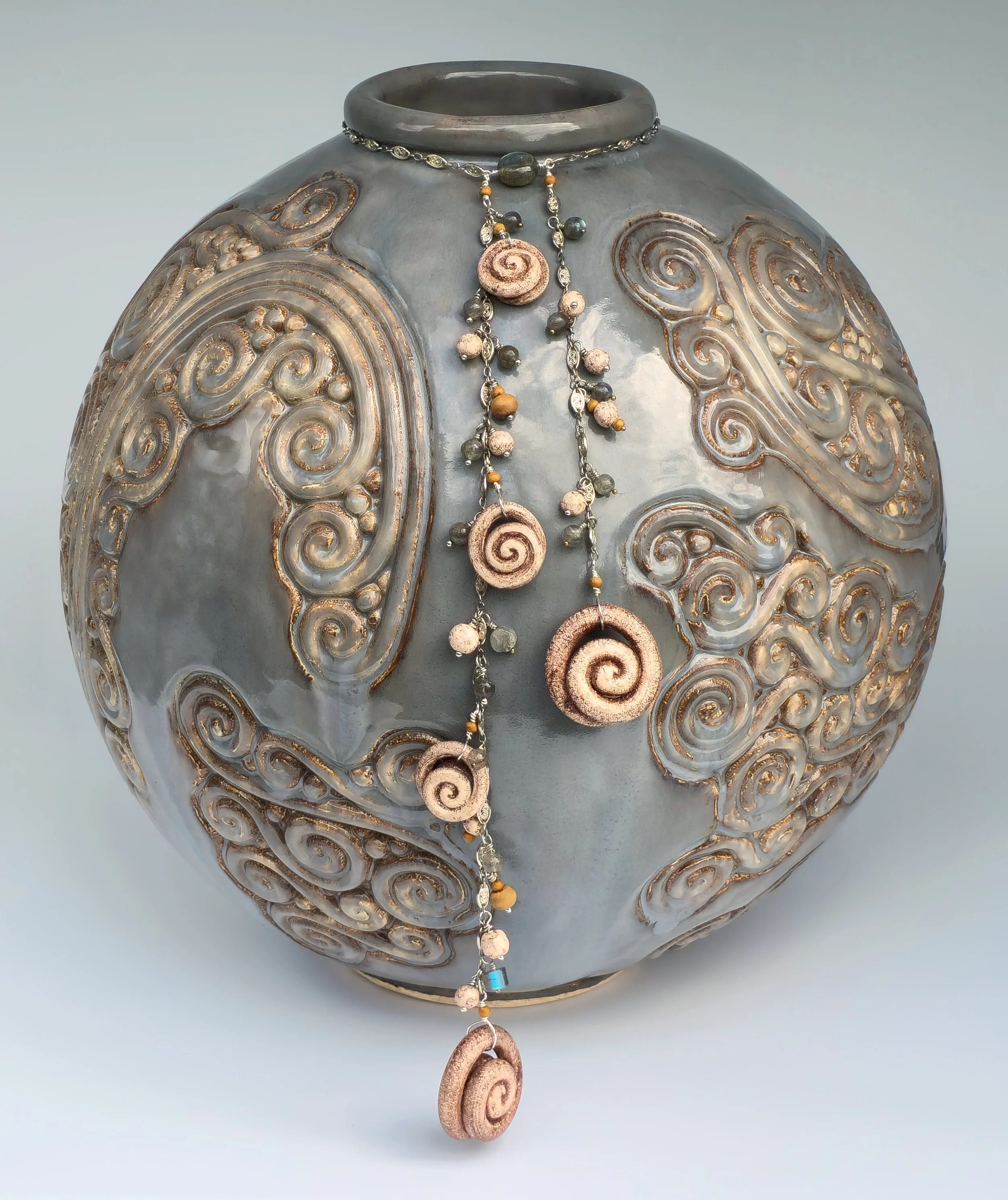 A round ceramic vase with a grayish glaze and gold-colored swirling designs. It has a layered, textured surface. A necklace with seashell-shaped pendants, beads, and small decorative elements hangs around its neck.