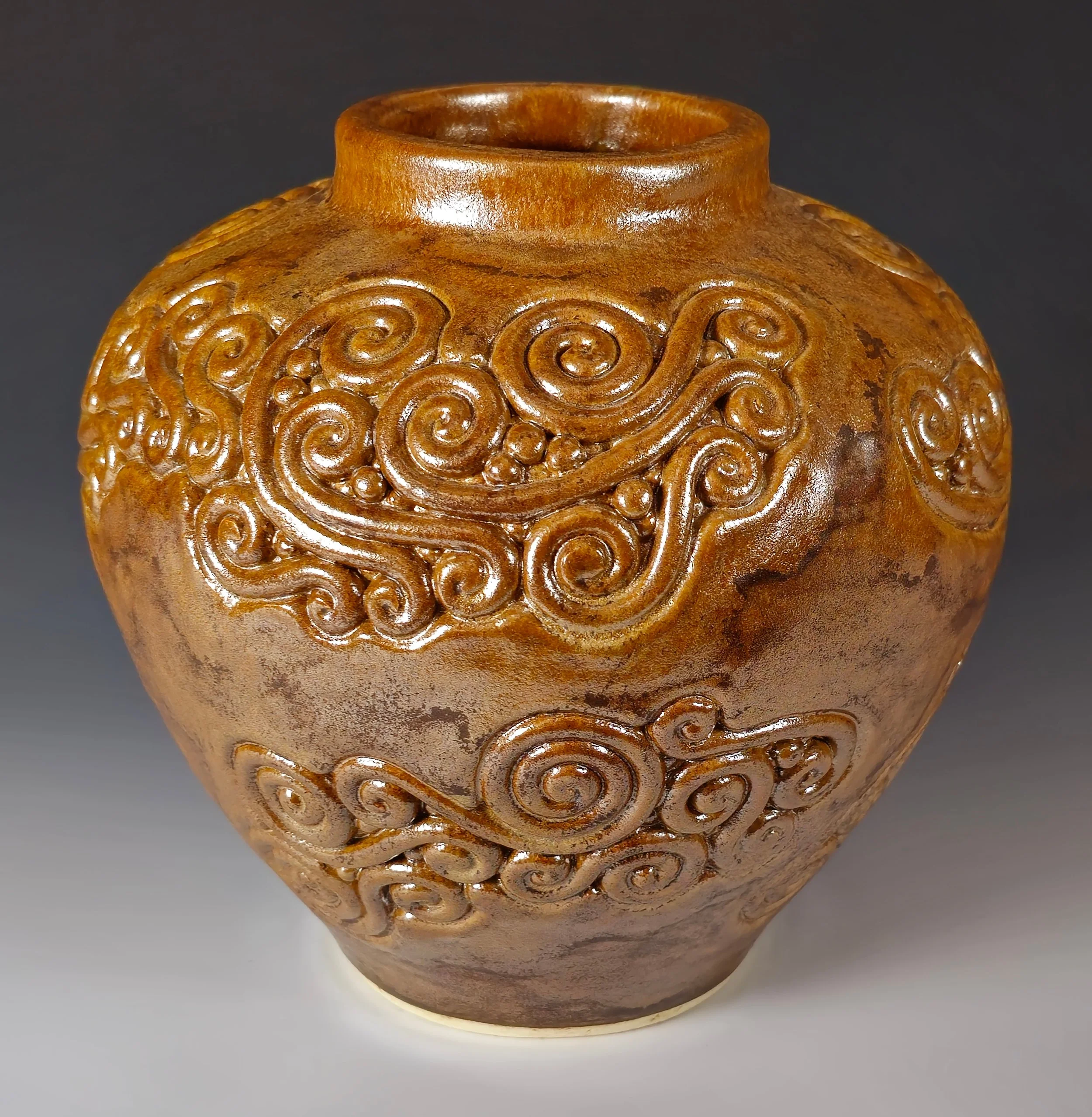 Close-up of a round, brown ceramic vessel with intricate swirl and dot carvings on the upper section, against a dark background.
