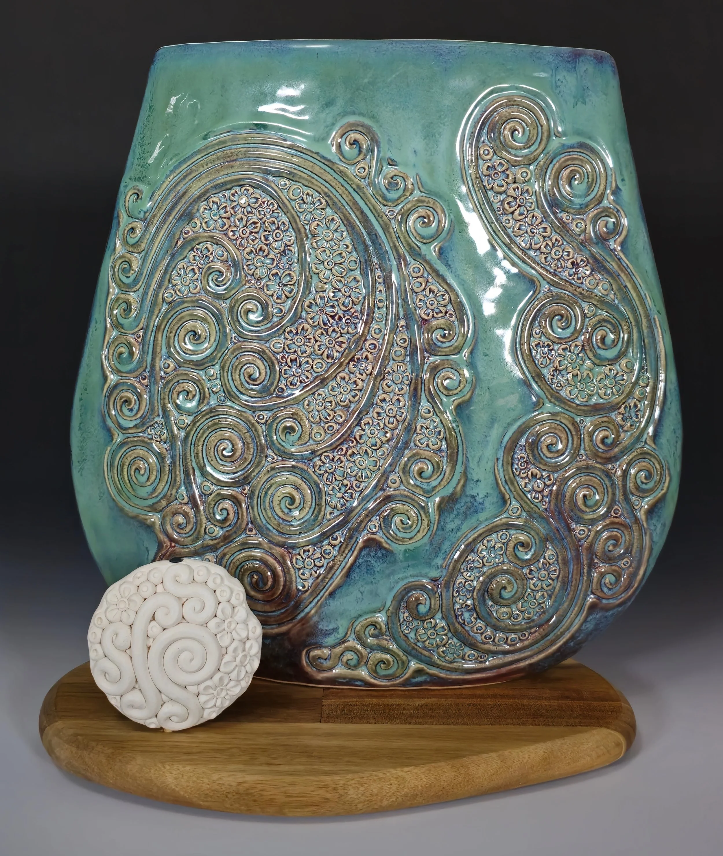 Ceramic vase with intricate swirling floral patterns in gold and blue over a gradient green glaze, mounted on a wooden base with a white carved decorative object in front.