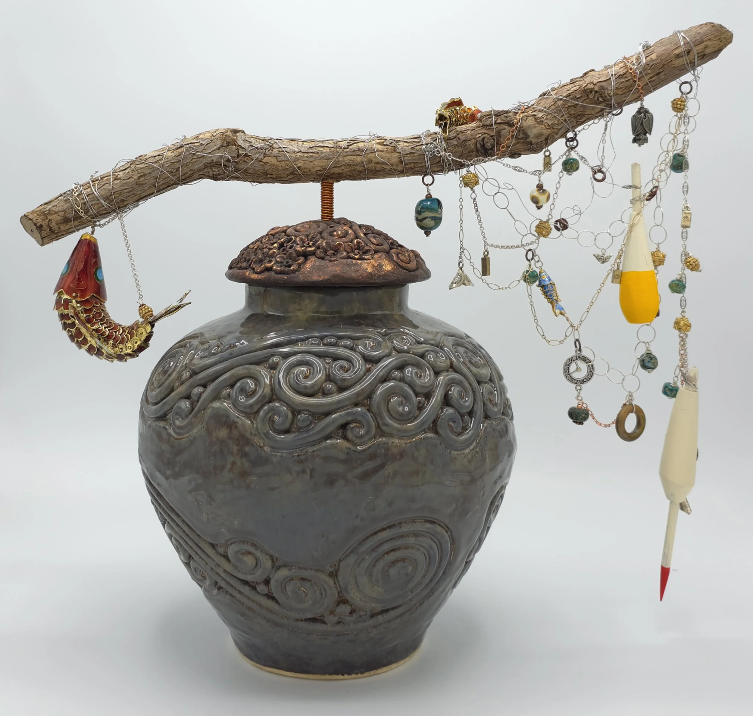 A decorated ceramic vase with a wooden branch draped with various jewelry items, including necklaces and pendants, resting on a white surface against a plain light background.