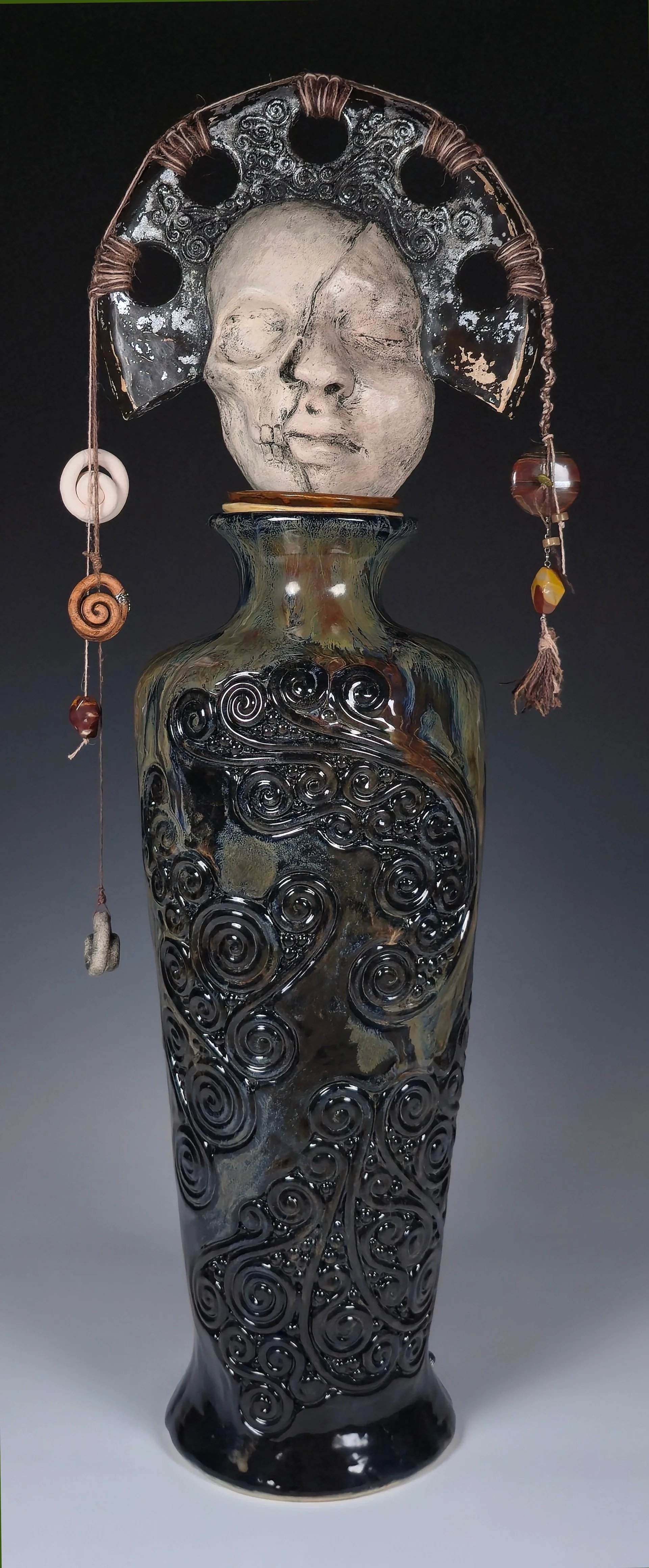An artistic glass sculpture of a female face with closed eyes, adorned with a decorative headpiece and hanging beads, mounted on a tall, intricately designed glass vase.