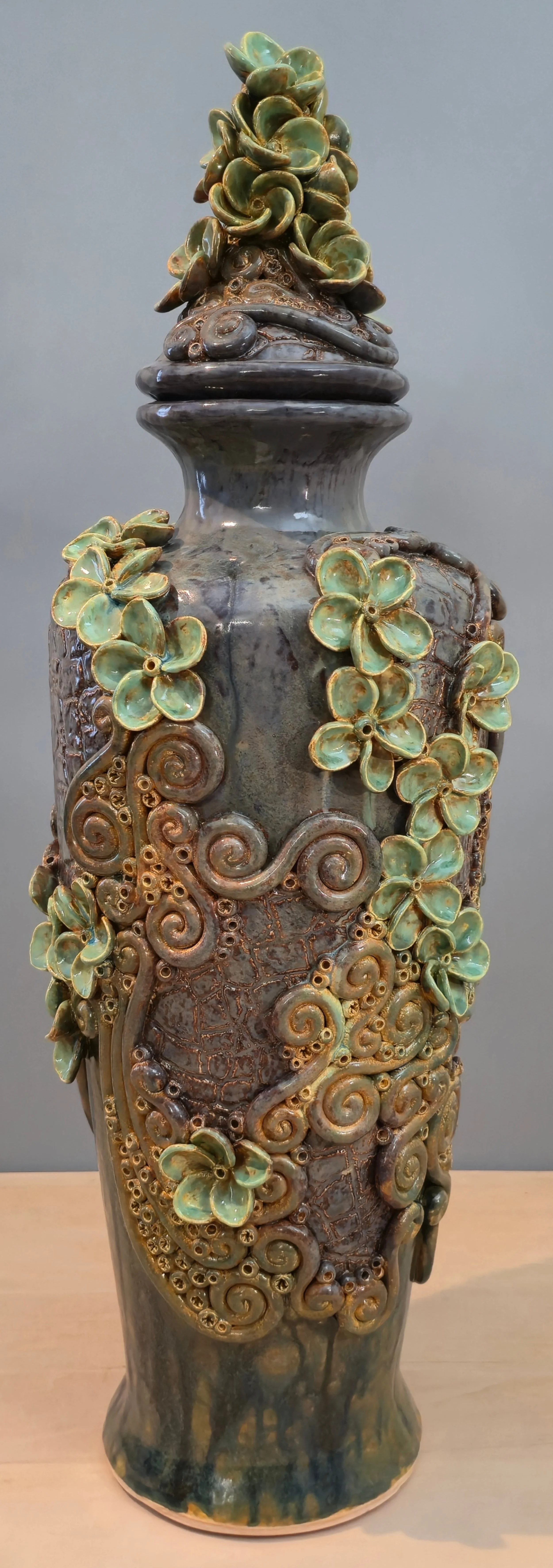 Ceramic vase with intricate swirling vine and flower design, featuring green and brown hues, with a textured surface and shiny glaze.