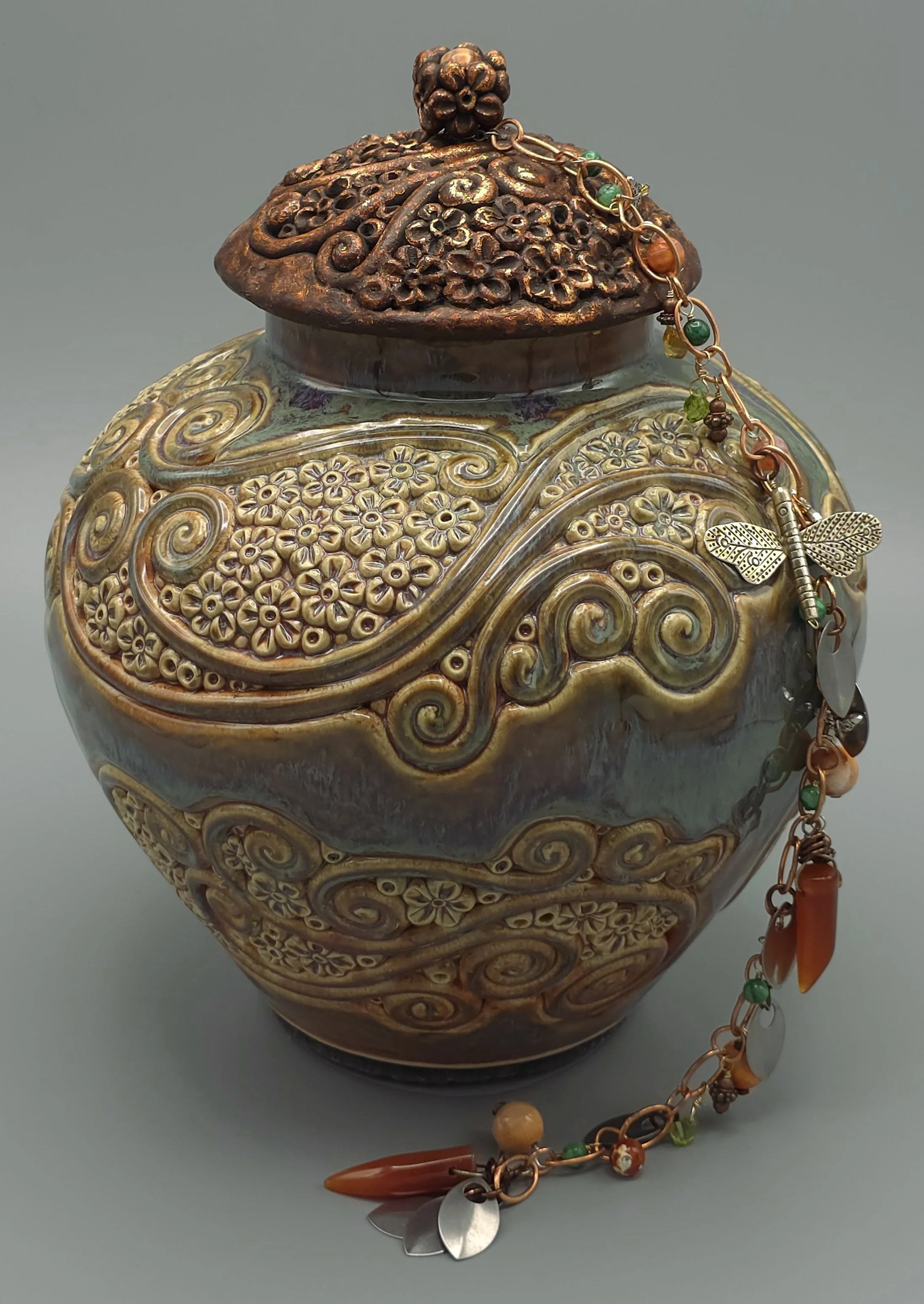 A decorative vintage-style ceramic vase with intricate floral and swirling patterns, topped with a carved wooden lid. A beaded chain with a metallic butterfly charm and various colorful beads and pendants is draped over the vase.