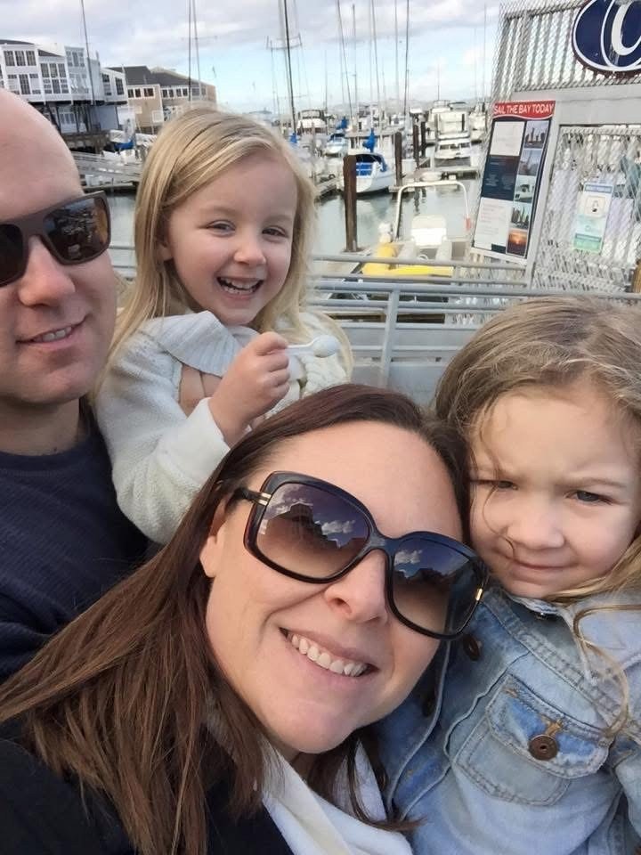 A happy family of four taking a selfie at a marina with boats and docks in the background. The woman is wearing sunglasses, and two girls are smiling while one appears shy or upset.