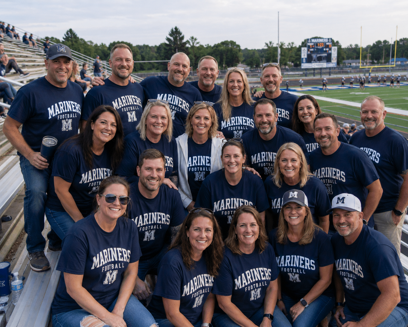 Group of people wearing navy blue 'Mariners Football' T-shirts posing for a photo at a football stadium with bleachers and a game in progress in the background.