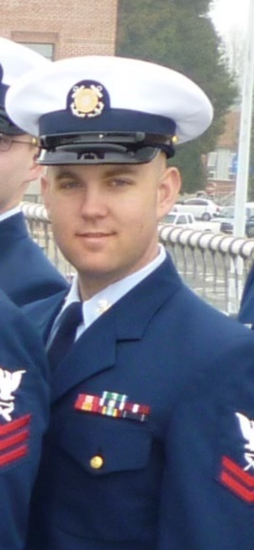A young man in a United States Air Force uniform with a white officer's cap, ribbons, and badges, standing outdoors near a fence with buildings and trees in the background.