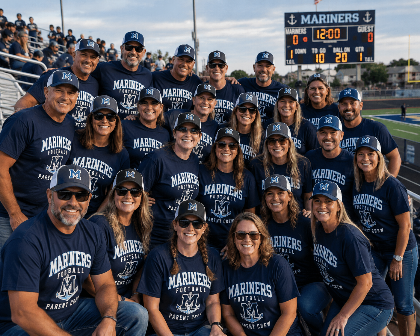 Group of people wearing matching Mariners Football Parent Crew shirts and caps on a football field with a scoreboard and stadium stands in the background.