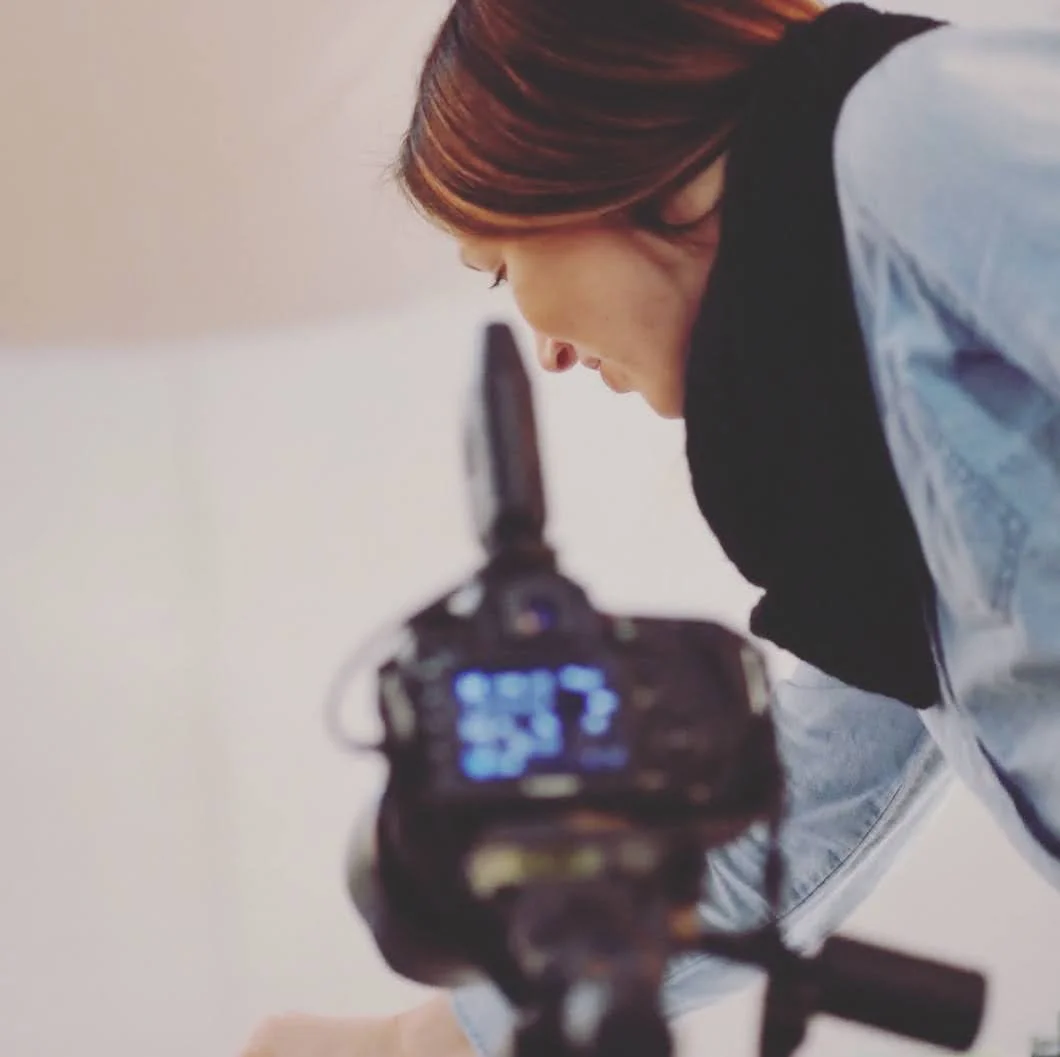 A woman with reddish-brown hair is leaning forward, looking at a camera or electronic device on a tripod.