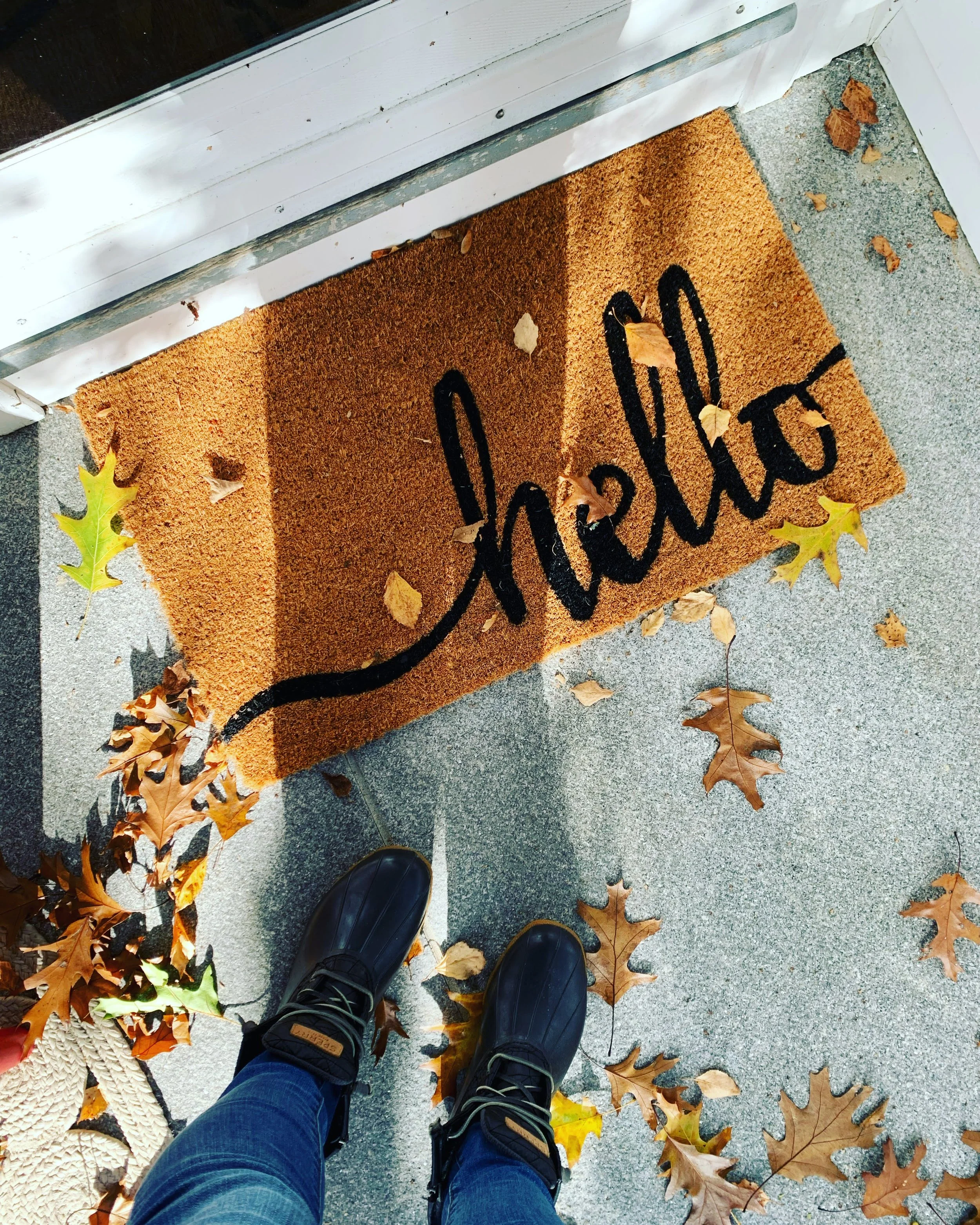 Person standing on a concrete porch with a doormat reading 'welcome,' surrounded by fallen autumn leaves, with black shoes and denim jeans visible.