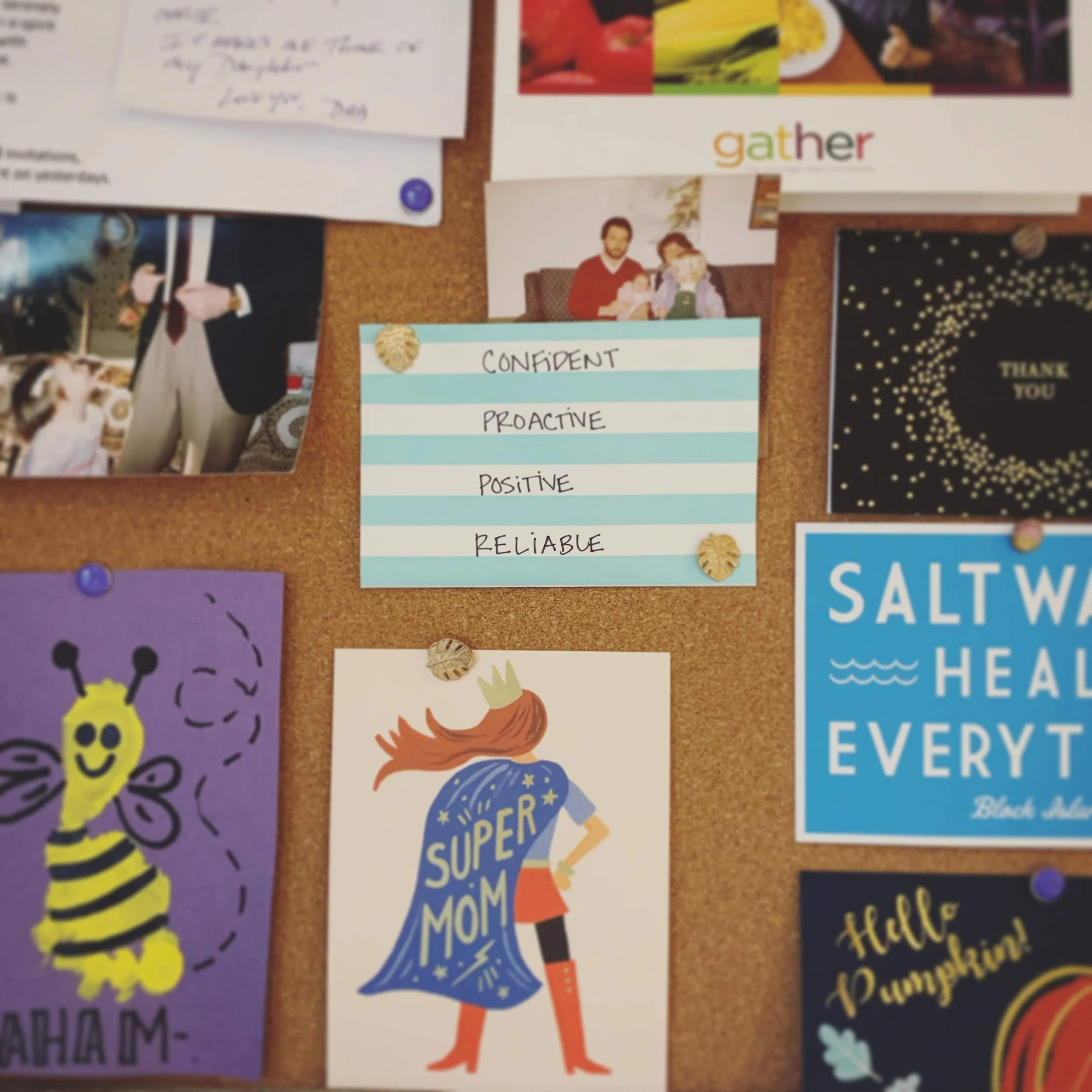 Colorful bulletin board with various photos, notes, and motivational words. A handwritten note with 'Love you, Dad' is visible. A word 'gather' is printed in colorful letters. A card shows a woman and a child with Christmas-themed text, and there is a purple card with a yellow cartoon character.