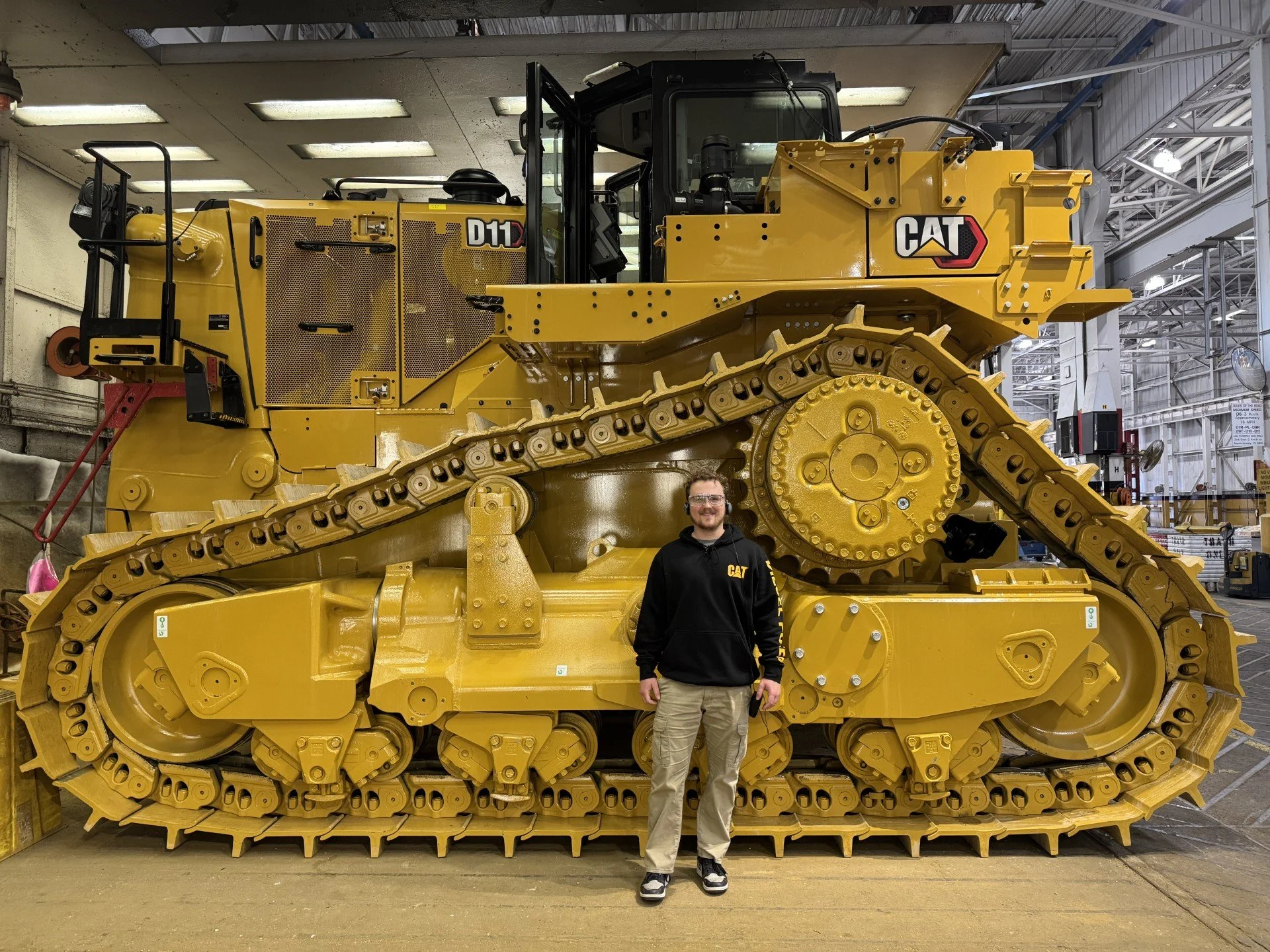 A person standing in front of a large yellow Caterpillar bulldozer inside a warehouse or factory. The person is smiling and wearing glasses, a black jacket with a CAT logo, beige pants, and sneakers.