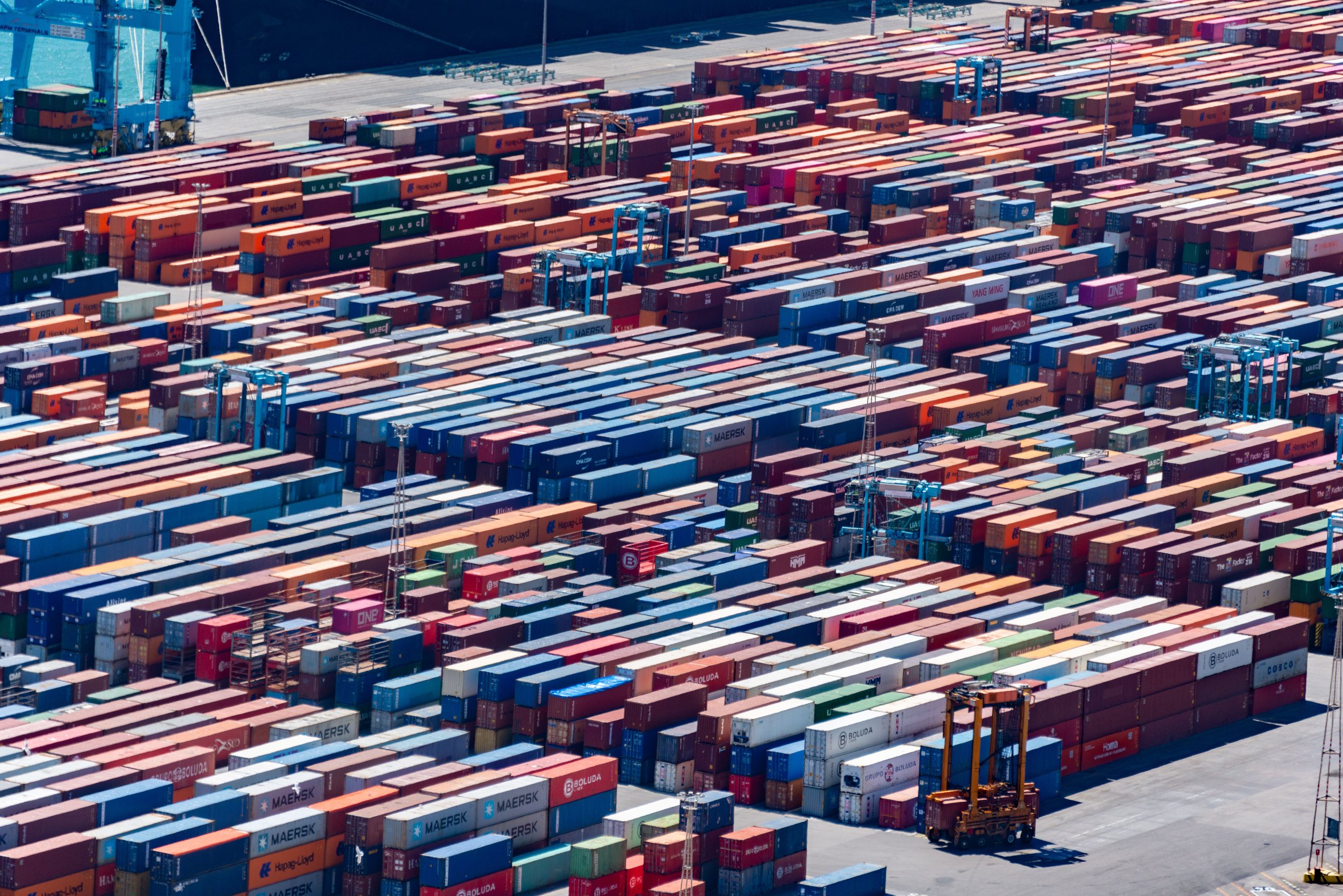 Many stacked cargo shipping containers in a port, with cranes and a cargo ship in the background.