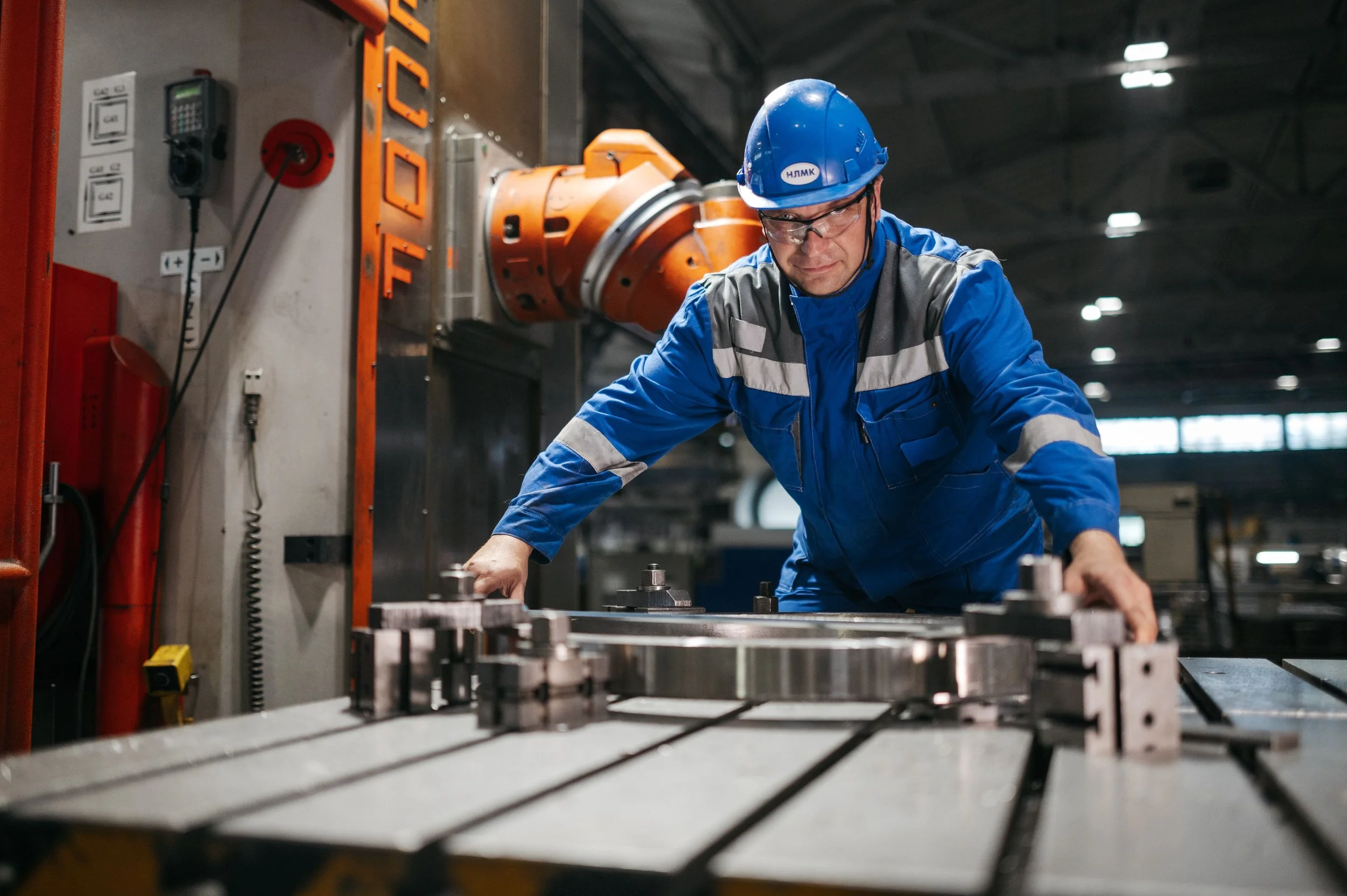 A factory worker in blue protective gear, including a hard hat and safety goggles, is working on machinery in an industrial setting with metallic components and equipment.