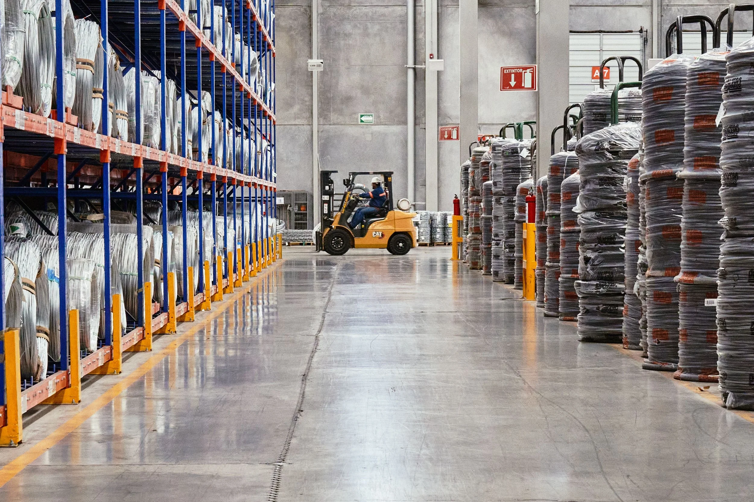 Inside a warehouse, a worker operates a forklift between shelves filled with coiled cables and stacks of wrapped pallets.