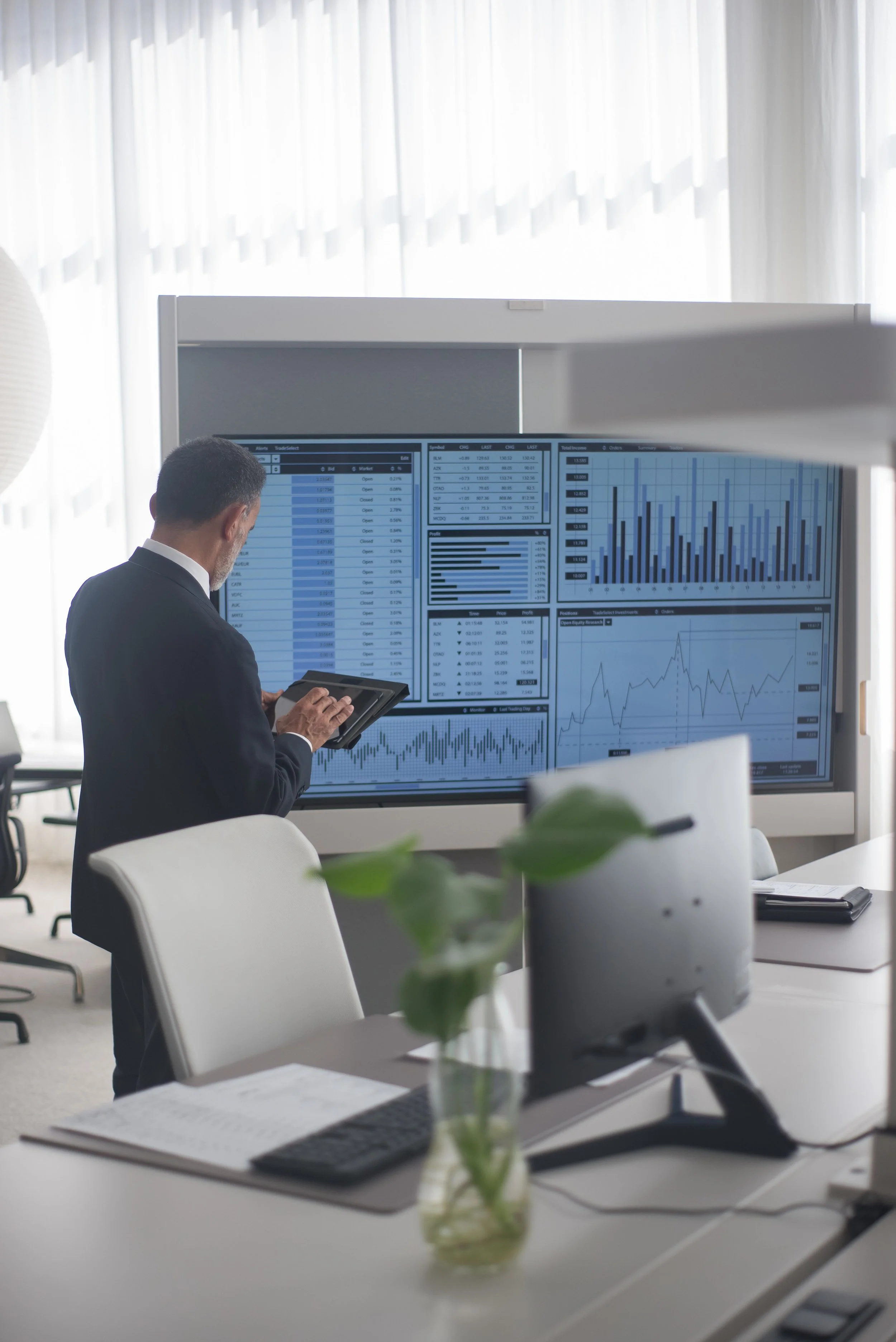 A man in a suit working on a tablet in an office with large screens displaying financial charts and data.