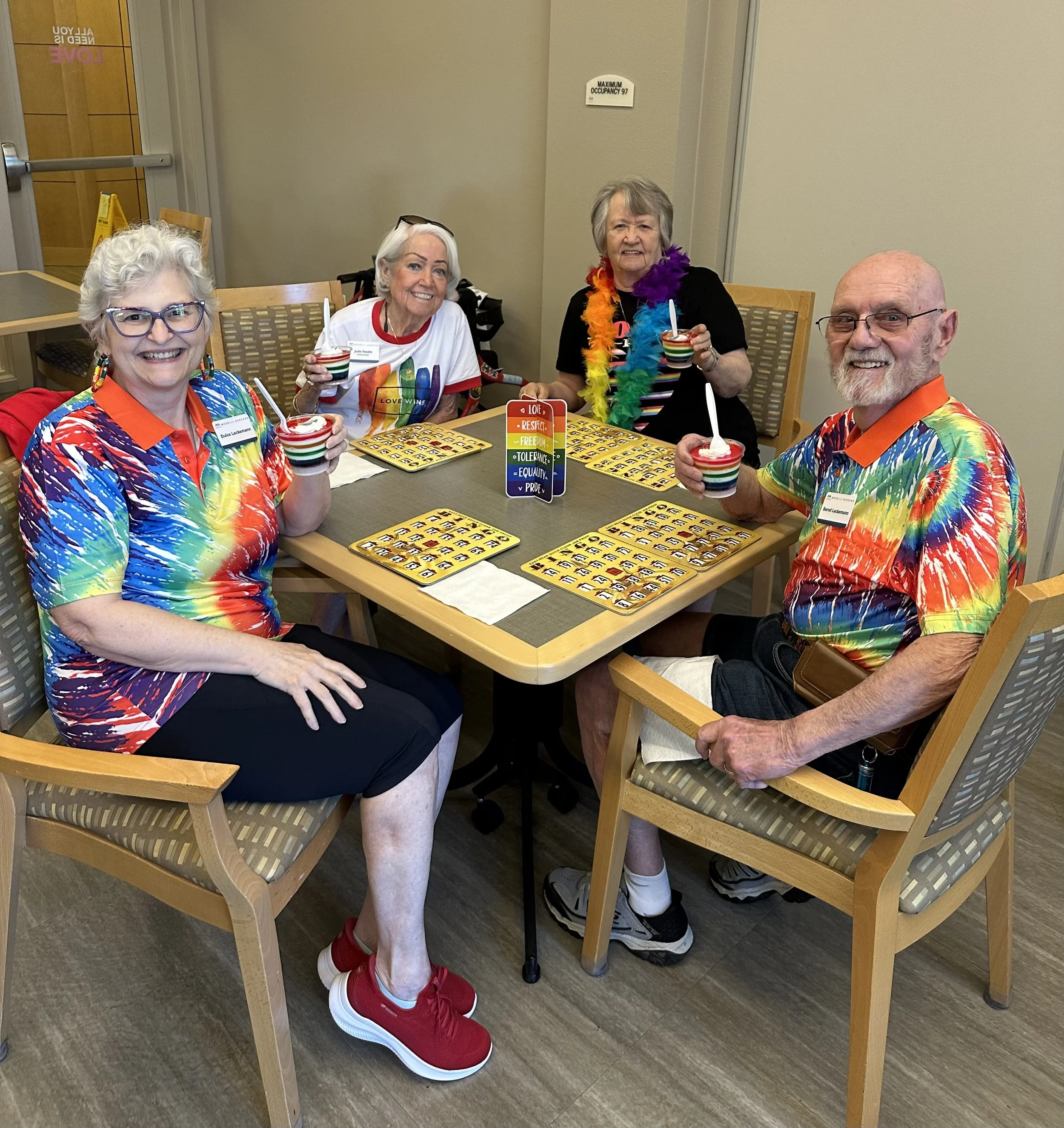Four smiling seniors sitting at a table in a room, holding colorful striped cups with white spoons, wearing rainbow-colored clothing and accessories, with bingo cards on the table, in a cheerful party setting.