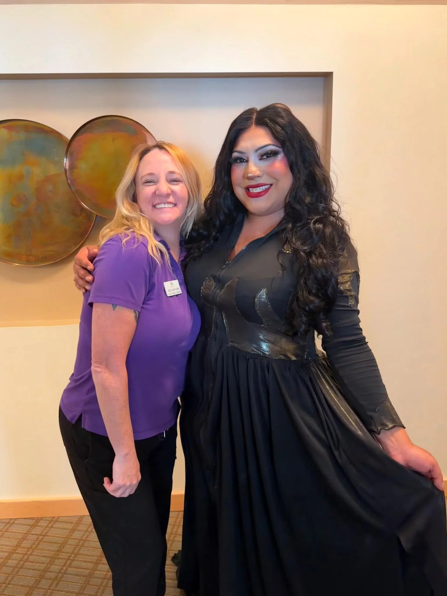 Two women standing close together, smiling, in an indoor setting with a beige wall and circular metal wall art. The woman on the left has blonde hair, wearing a purple short-sleeve top, and the woman on the right has long dark curly hair, wearing a black dress.
