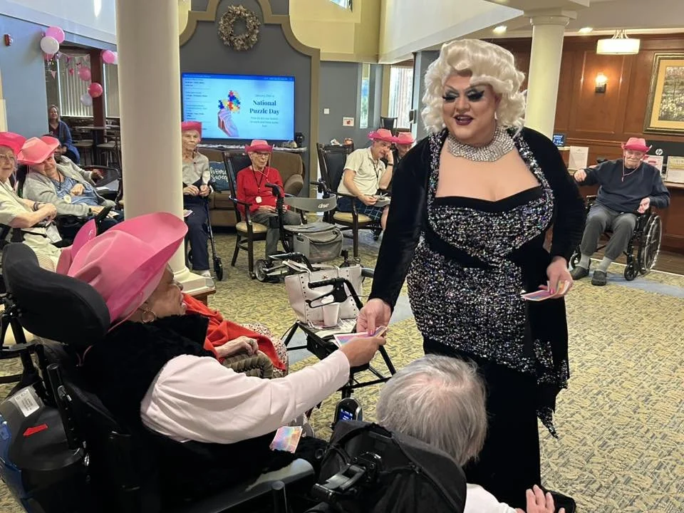 A drag queen performer in a sparkly dress and blonde wig interacts with elderly women wearing pink hats at a senior living center, during a celebration for National Puzzle Day.
