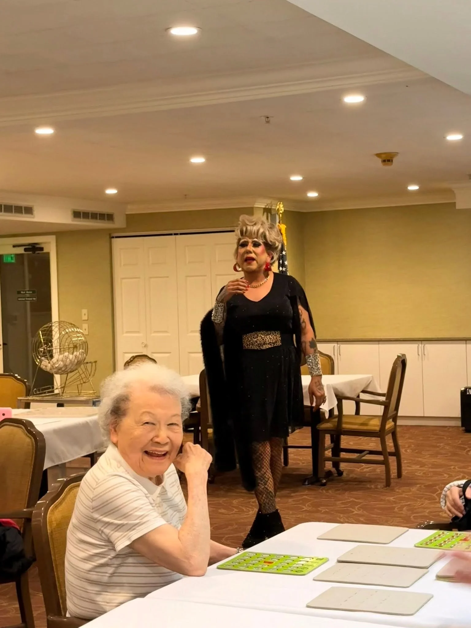 An elderly woman sitting at a table with her hand near her face, smiling, with a bingo game in front of her. In the background, a drag queen performer with gray hair, makeup, jewelry, and a black dress with fishnet stockings is standing and singing or talking. The setting appears to be a social event or gathering in a banquet hall.