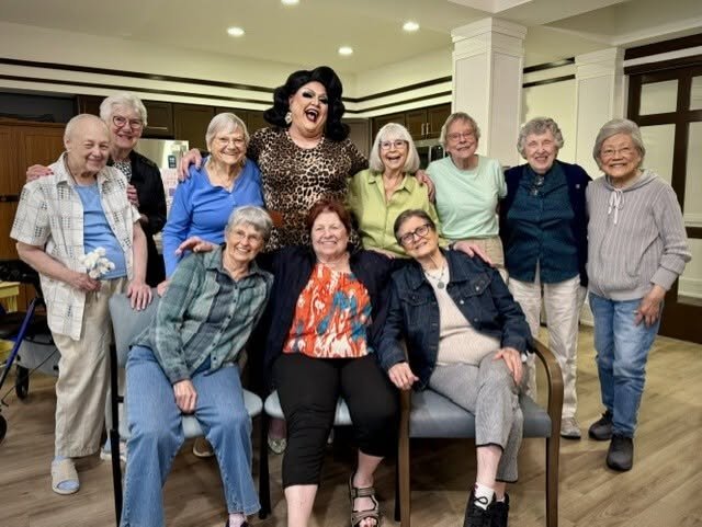 A diverse group older women smiling and posing together with a drag queen in a well-lit, senior living setting.