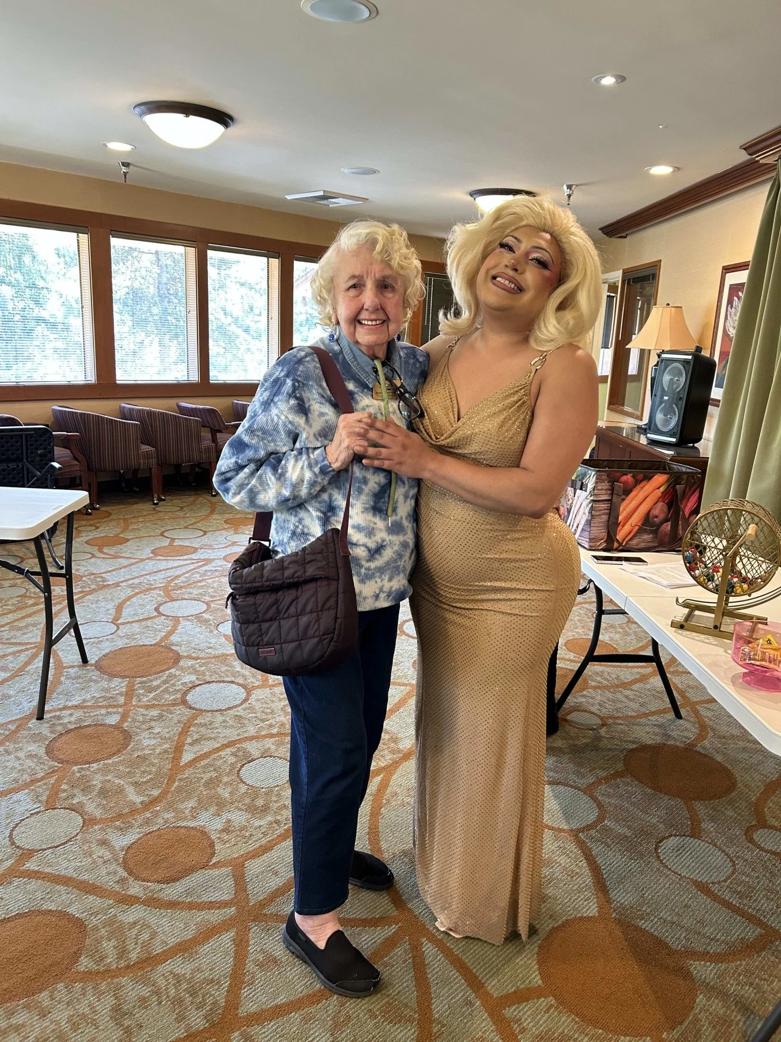 An older woman and a drag queen standing close together indoors, the older woman with curly blonde hair in a tie-dye jacket and glasses, and the drag queen with platinum blonde hair in a gold dress, smiling at the camera.