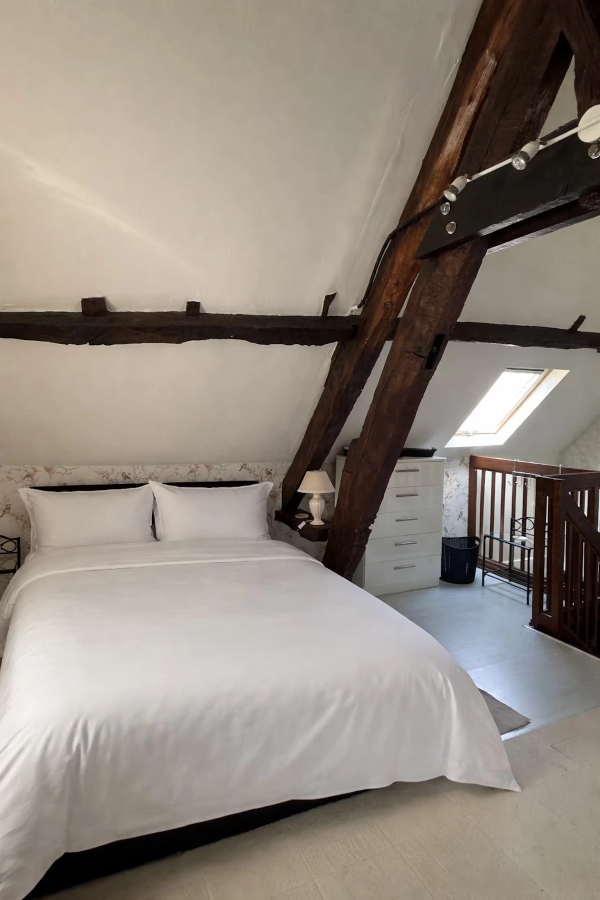 A bedroom with a white bed, a white dresser, a lamp, a skylight window, and exposed wooden beams on the ceiling.
