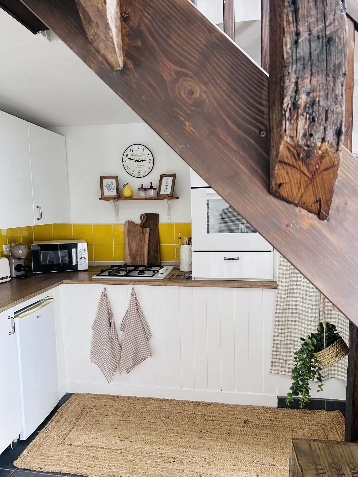 A cozy kitchen scene with a wooden countertop, white cabinets, a microwave, a small refrigerator, a stove, and a wall clock. Decor includes framed botanical prints, a yellow vase, and potted plants. There are checkered cloth towels hanging on the wall and a woven rug on the floor.