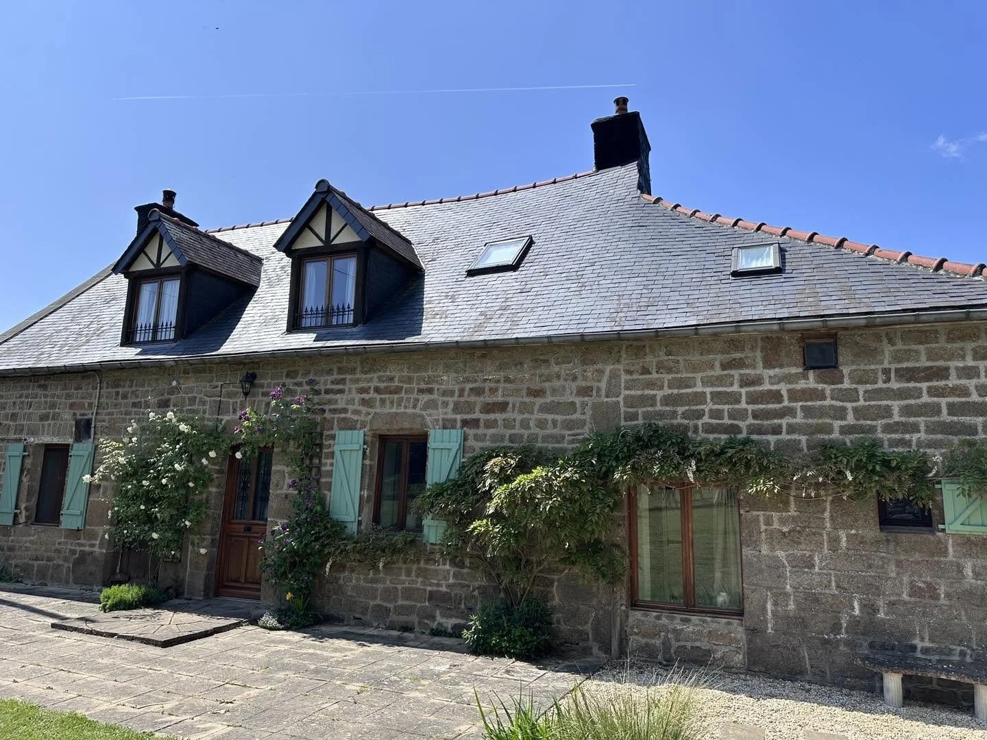 Le Tertre Farmhouse with green shutters, flowering bushes, a paved walkway, and a clear blue sky.