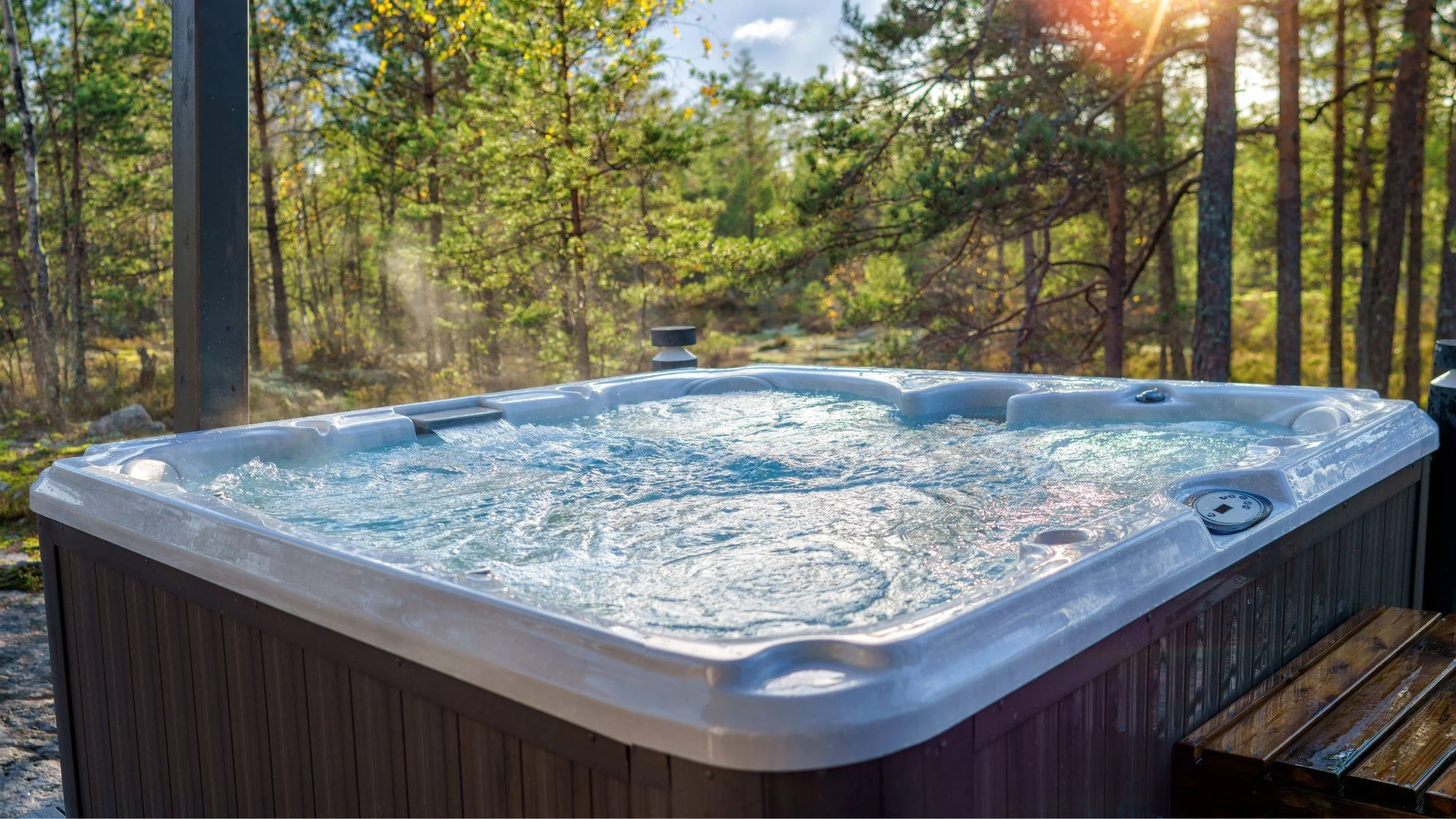 An outdoor hot tub filled with bubbling water, set in a wooded area with sunlight filtering through trees.