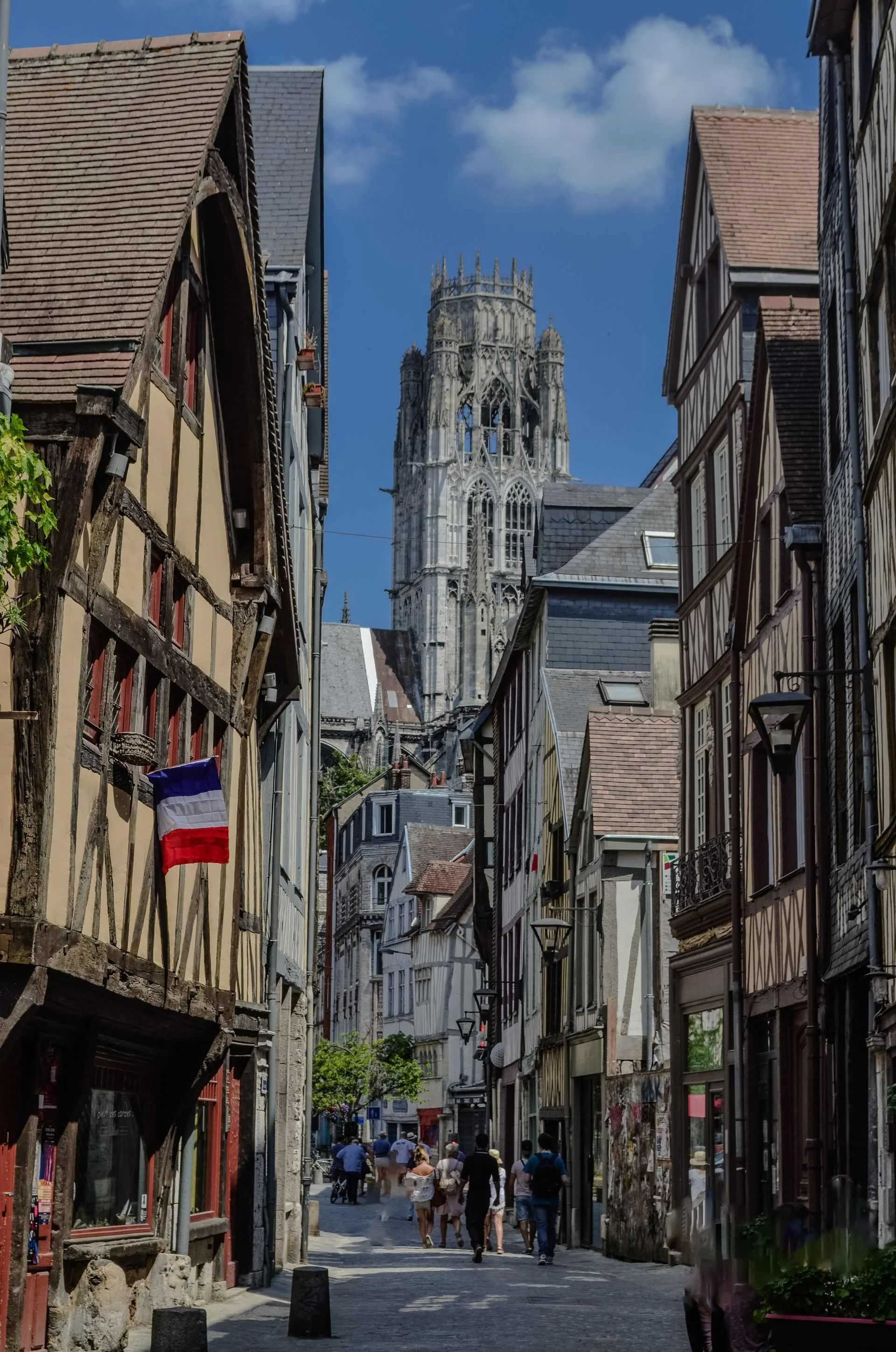 A narrow street in an old European city with half-timbered buildings on both sides. The street is busy with pedestrians and the distant Gothic cathedral tower is visible against a blue sky.